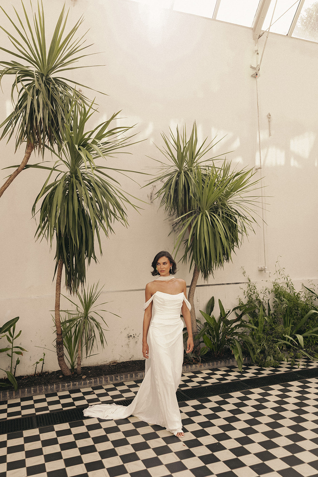 Woman in a ivory chiffon wedding dress standing in a room with plants and checkered floor.
