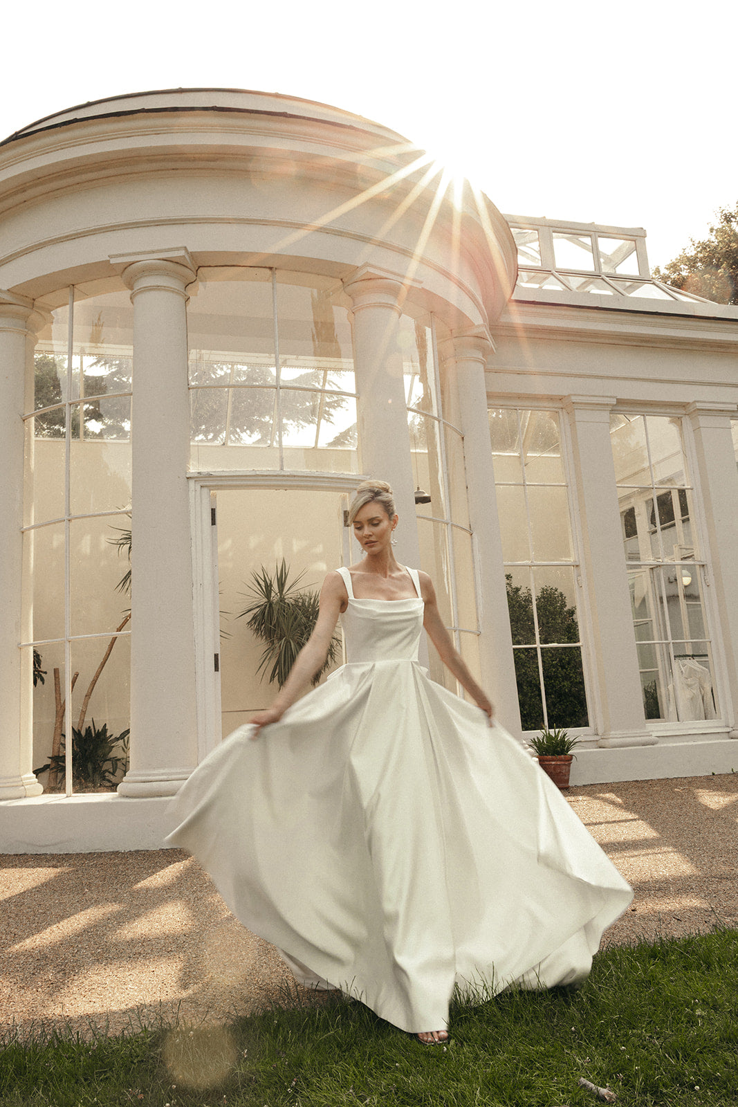 Woman in a ivory satin cowl neck wedding dress standing in front of a sunlit building with columns.