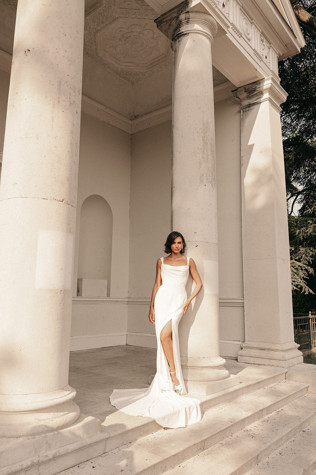 Woman in a ivory cowl neck wedding dress standing in front of classical architecture with columns.