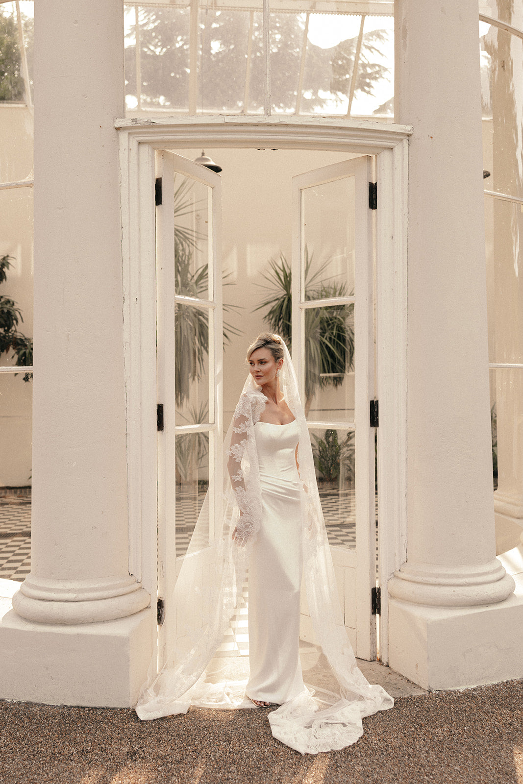 Woman in a ivory wedding dress and long lace wedding veil standing in front of a large glass door with columns.