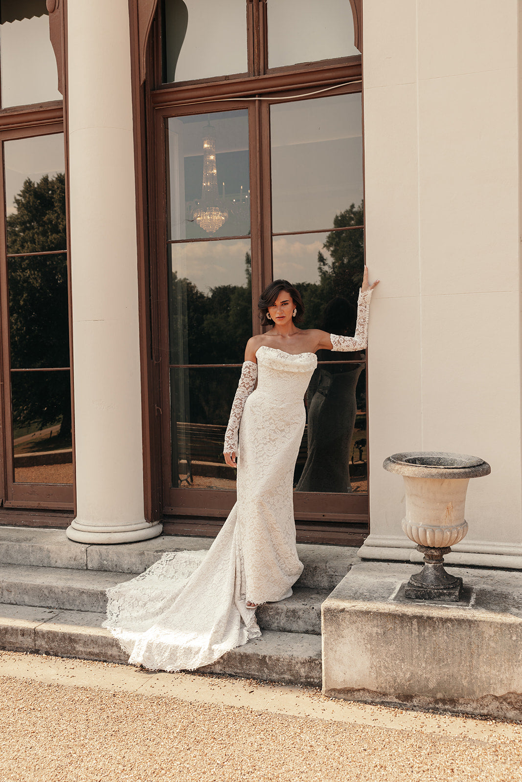 Woman in a ivory lace wedding dress standing on steps outside a building with large windows.