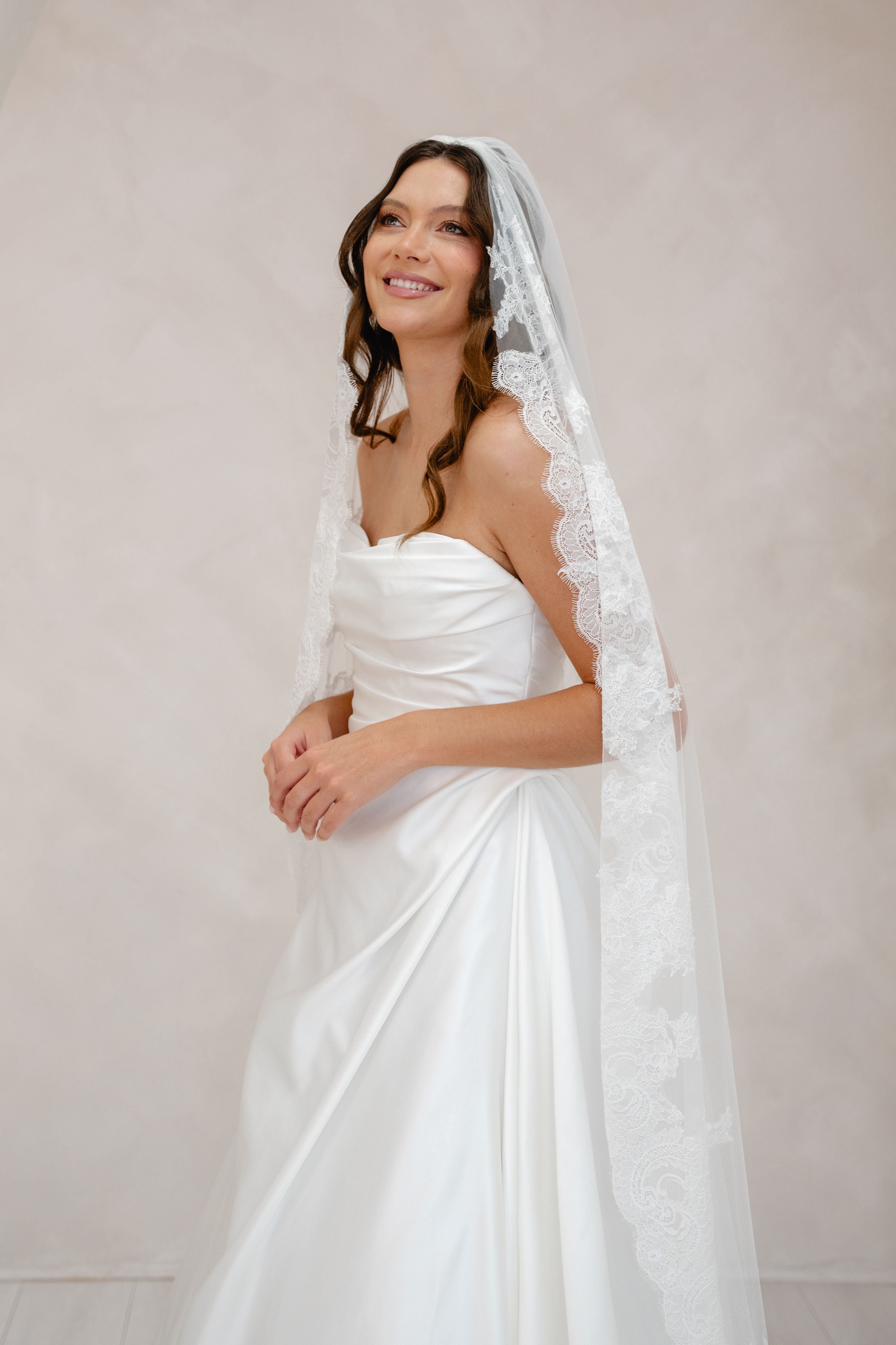 Woman wearing a white wedding dress with a lace veil against a plain background