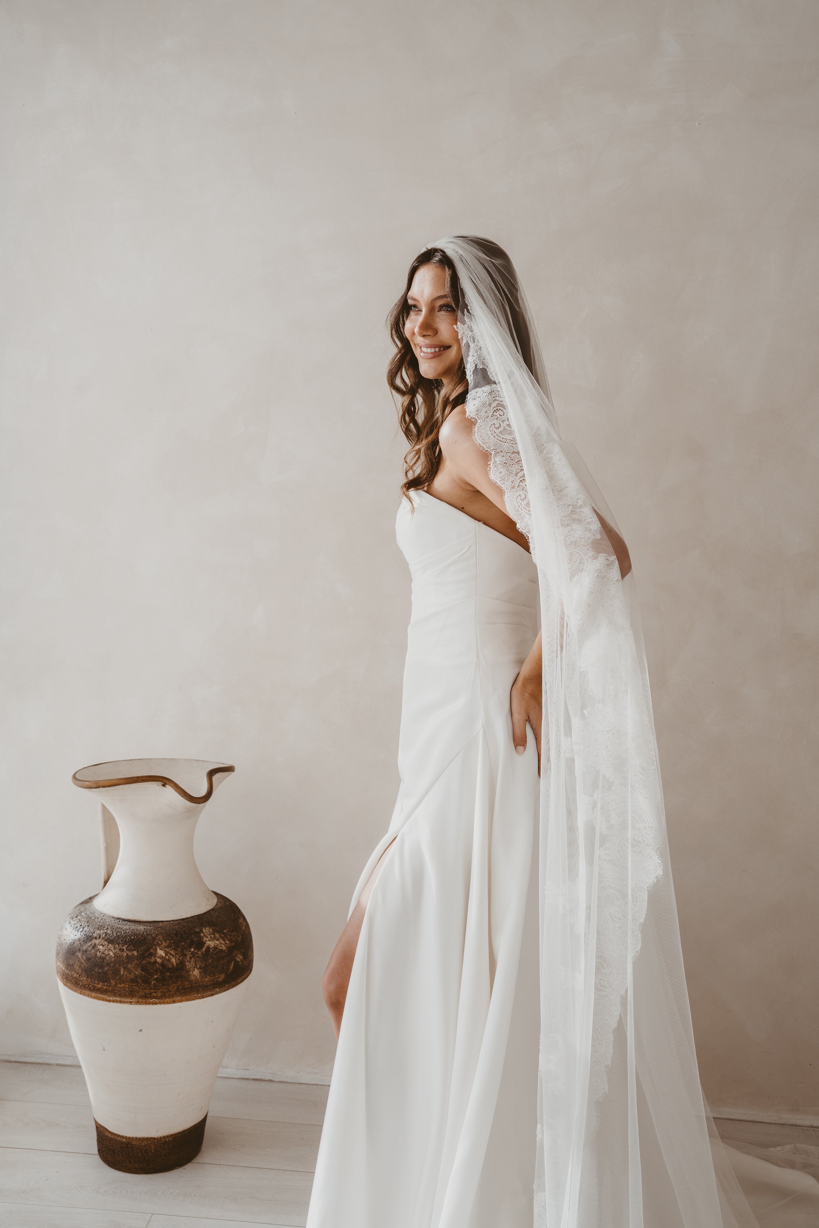 Woman in a white wedding dress with a veil standing next to a decorative vase against a plain wall.