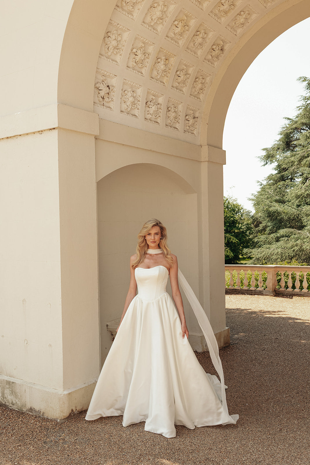 Woman in a ivory satin wedding dress standing under an archway with trees in the background