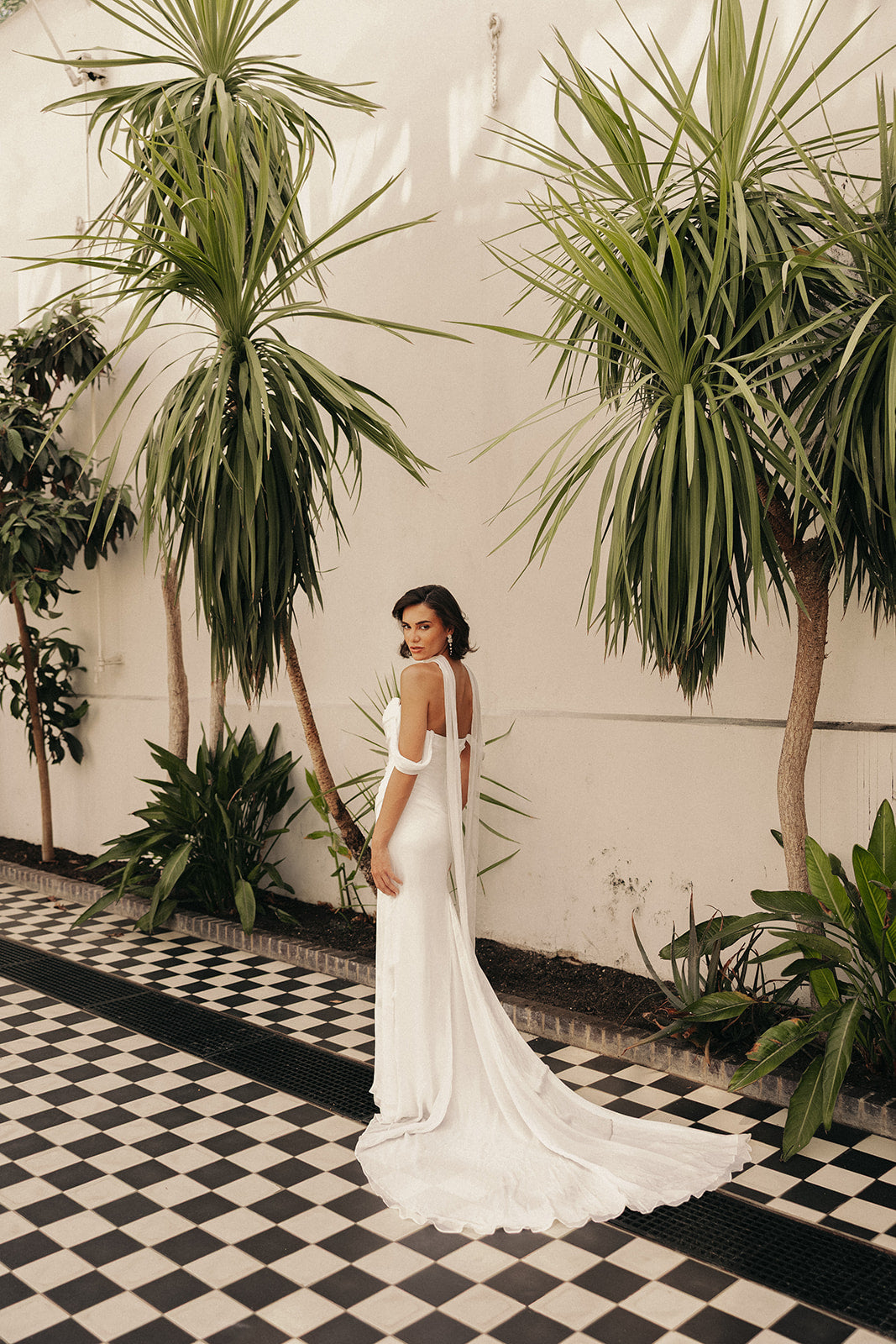 Woman in a ivory wedding dress standing in a tropical setting with plants and a checkered floor.