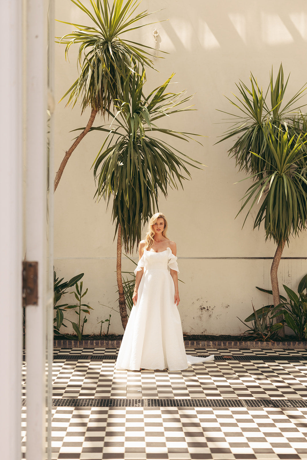 Woman in a ivory wedding dress standing in a sunlit room with plants and checkered floor.