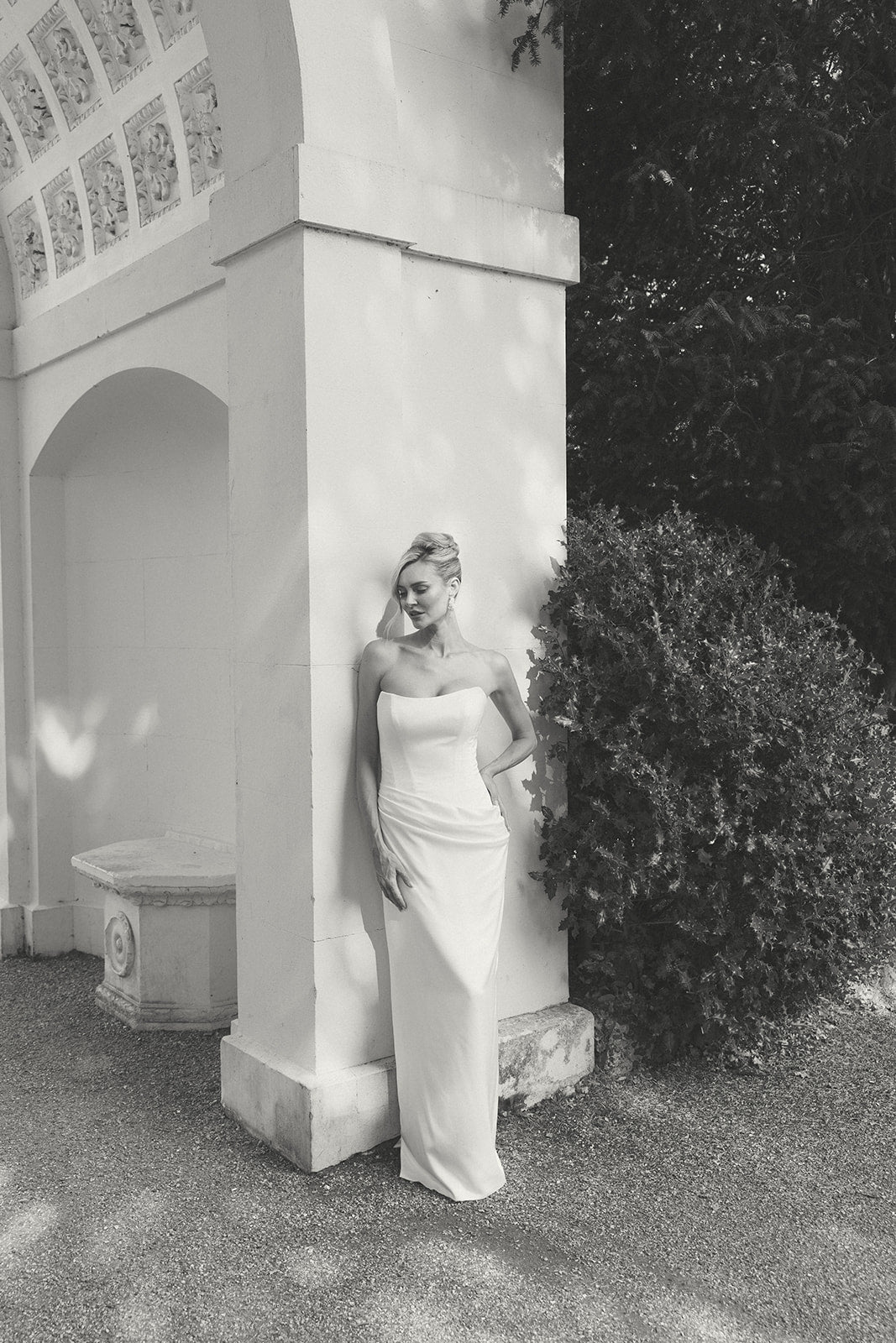 Woman in a strapless dropped waist wedding dress standing next to a classical architectural structure in a garden.