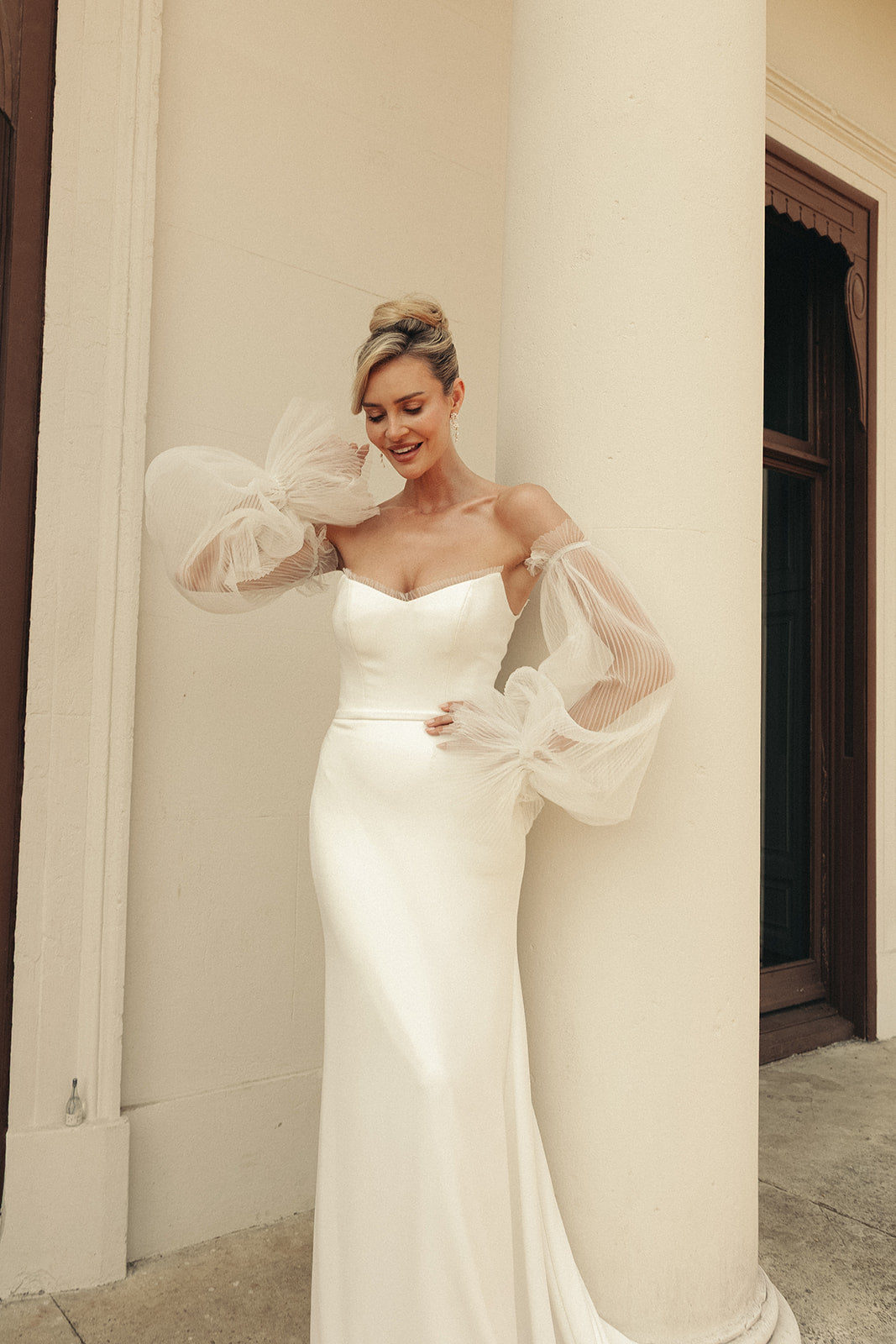 Woman in a ivory wedding dress with sheer sleeves standing against a classical building.