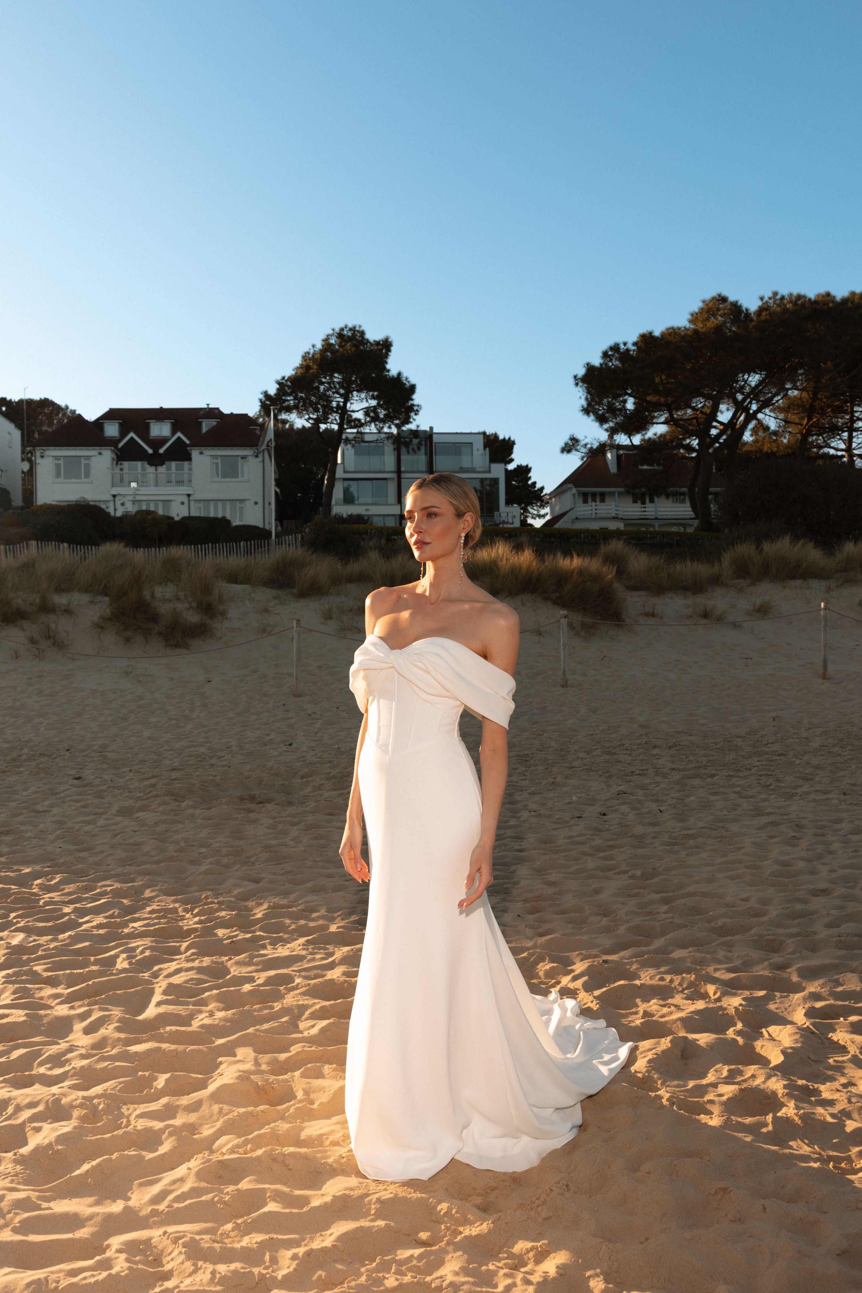 Woman in a white dress standing on a sandy beach with houses and trees in the background.