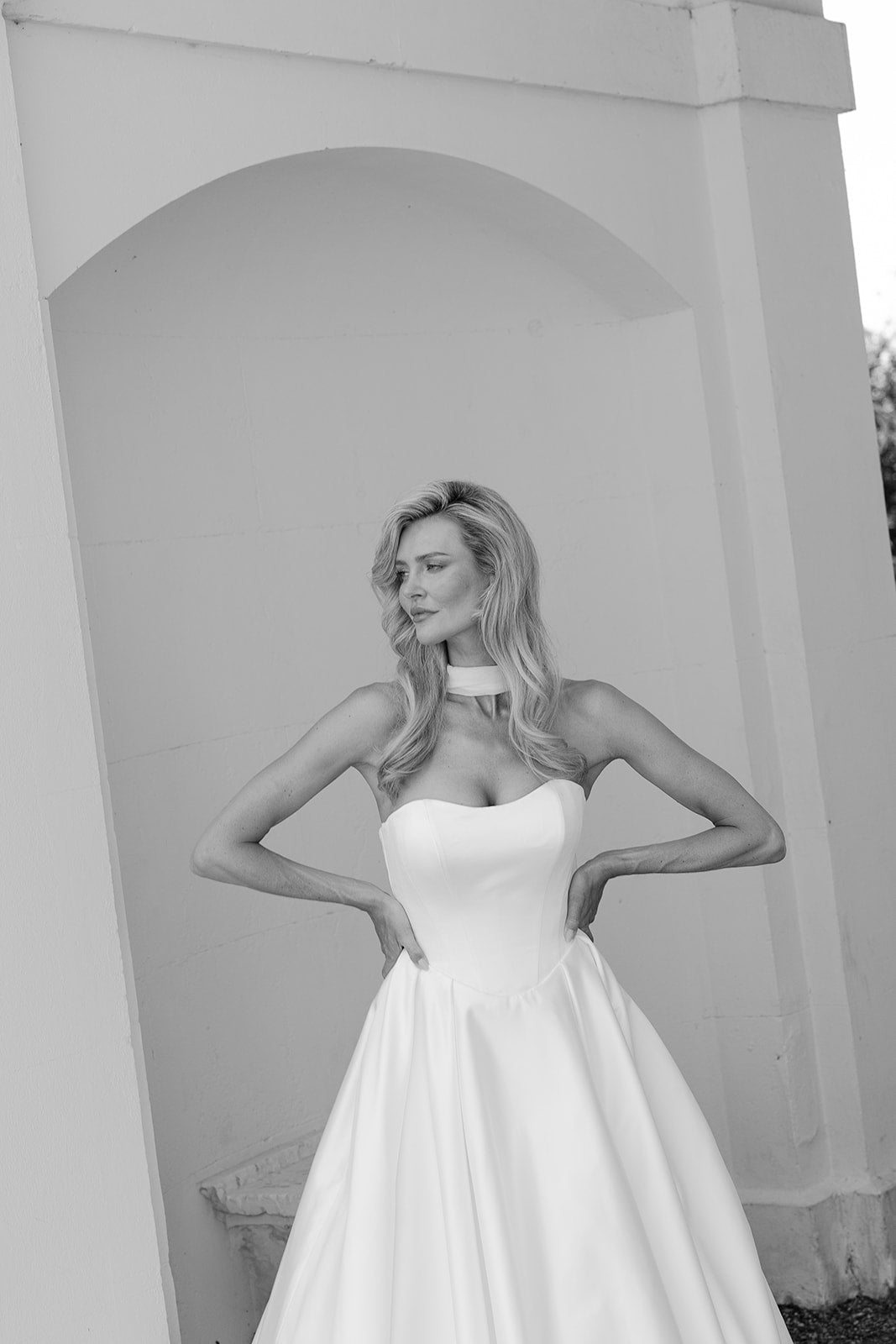 Woman in a strapless ivory satin wedding dress standing against a light-colored wall.