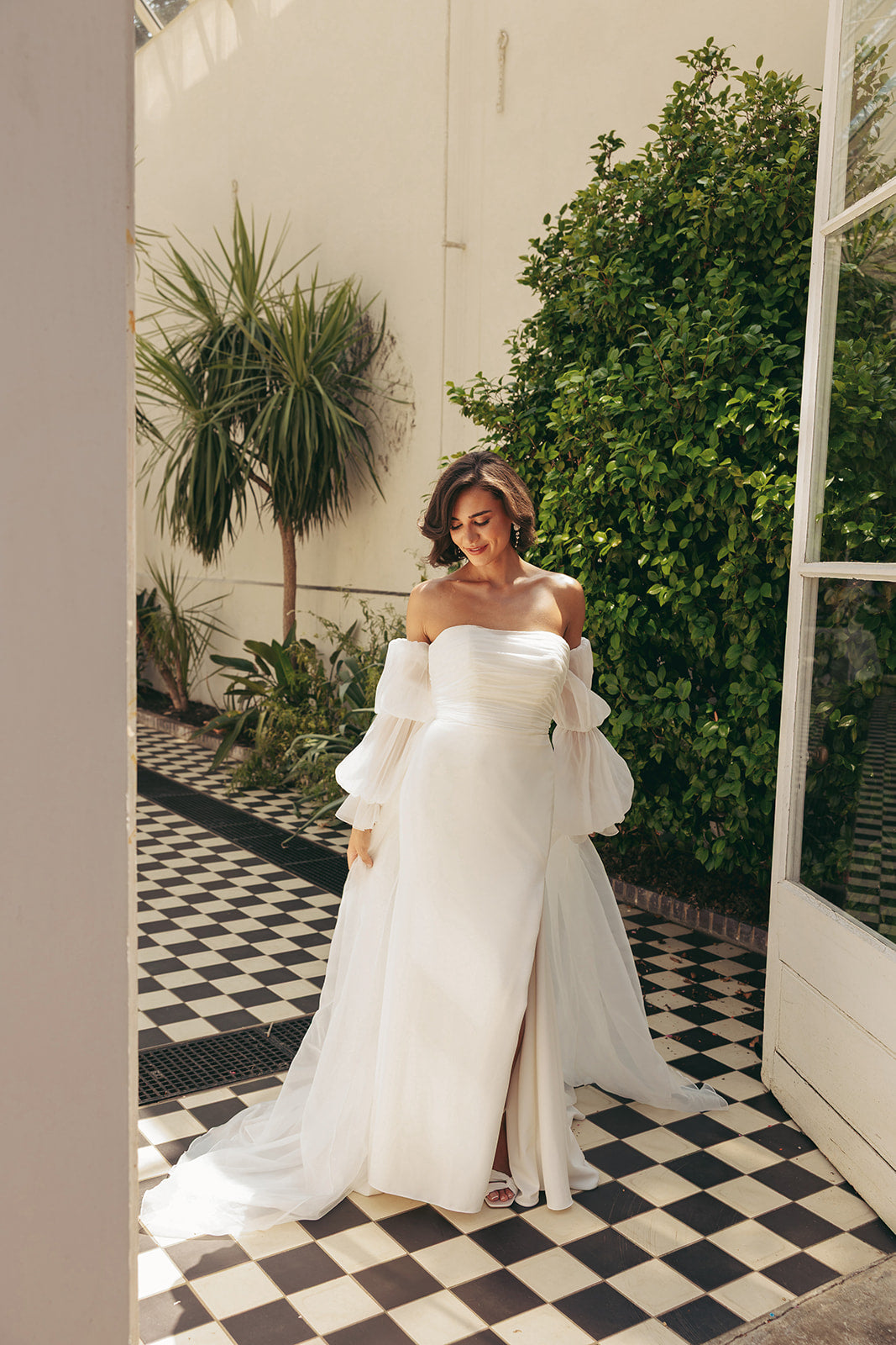 Woman in a ivory strapless wedding dress standing in a garden with plants and a checkered floor.