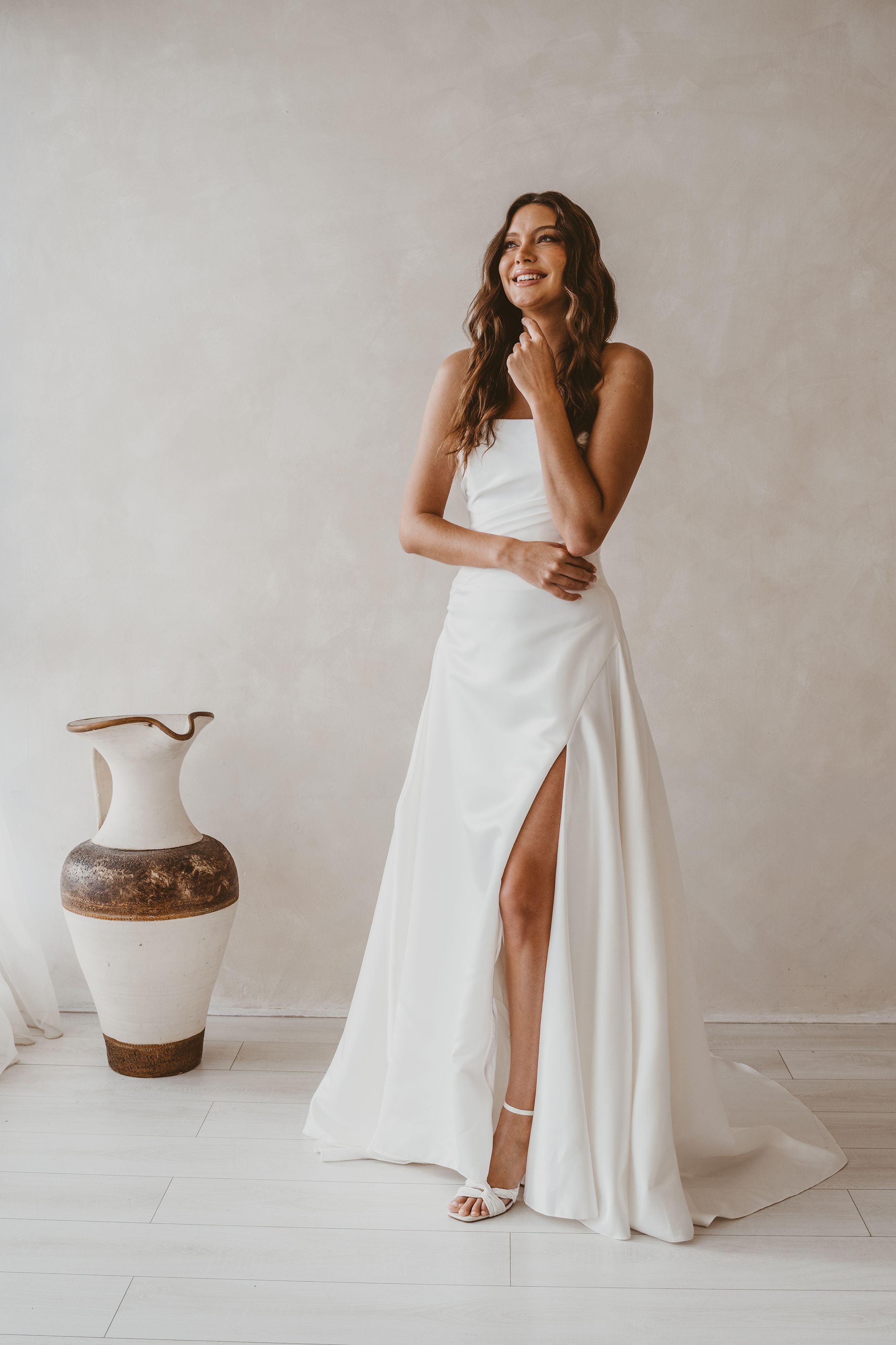 Woman in a white dress standing in a room with a vase and light-colored walls.