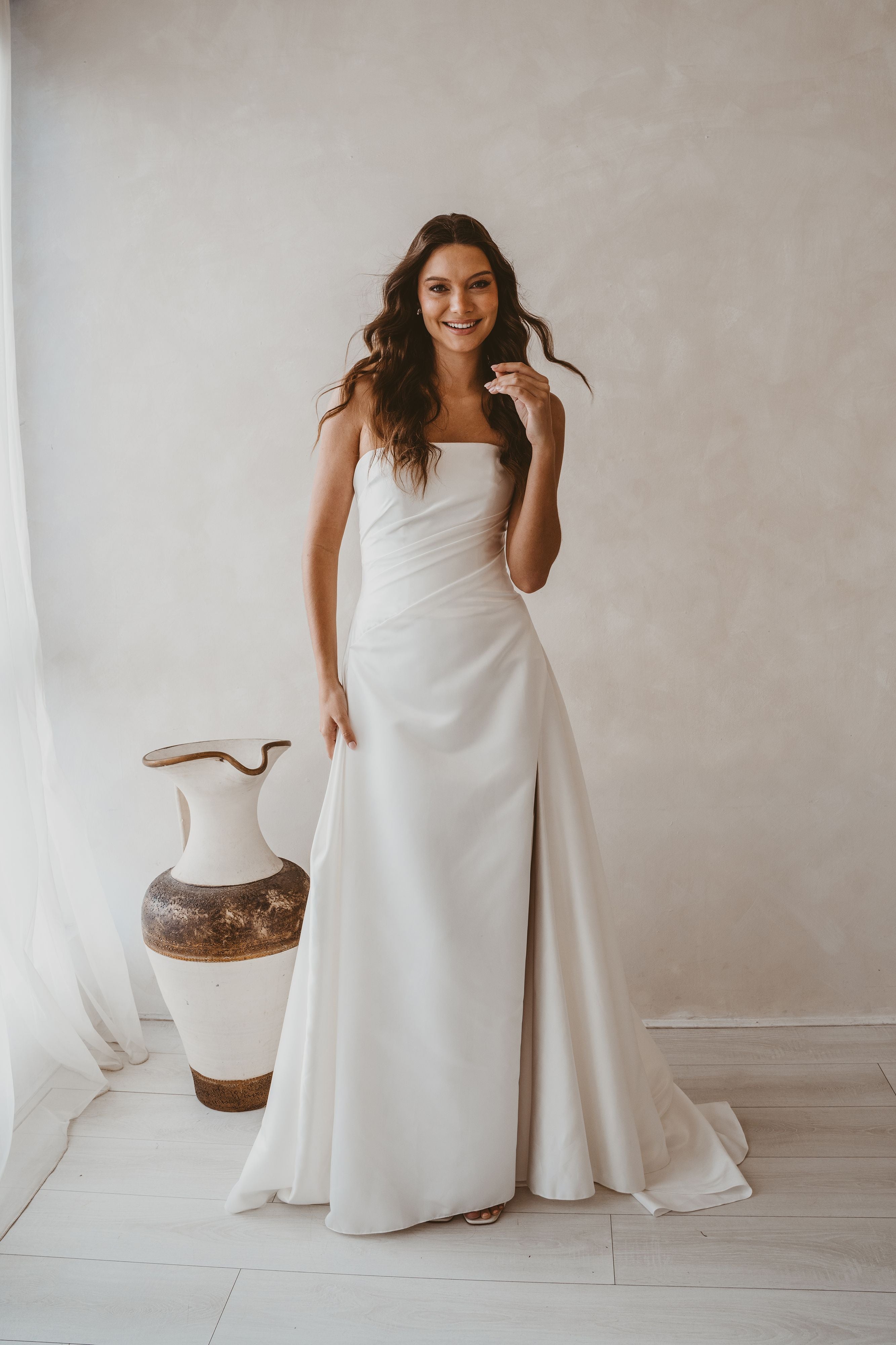 Woman in a white wedding dress standing next to a decorative vase against a plain wall.