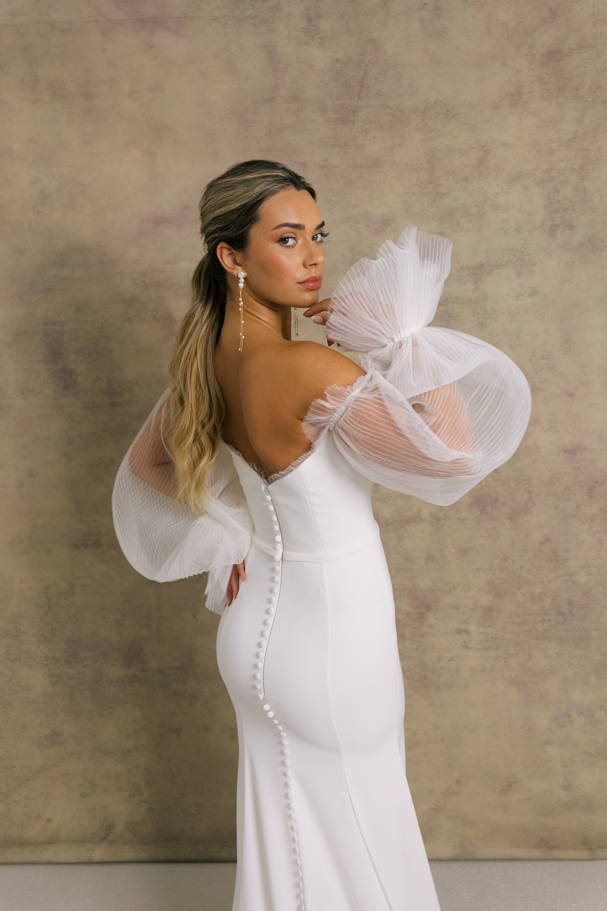 A woman in a strapless ivory wedding gown with a sweetheart neckline and sheer, pleated tulle sleeves showcasing the elegant fit and flare crepe skirt.