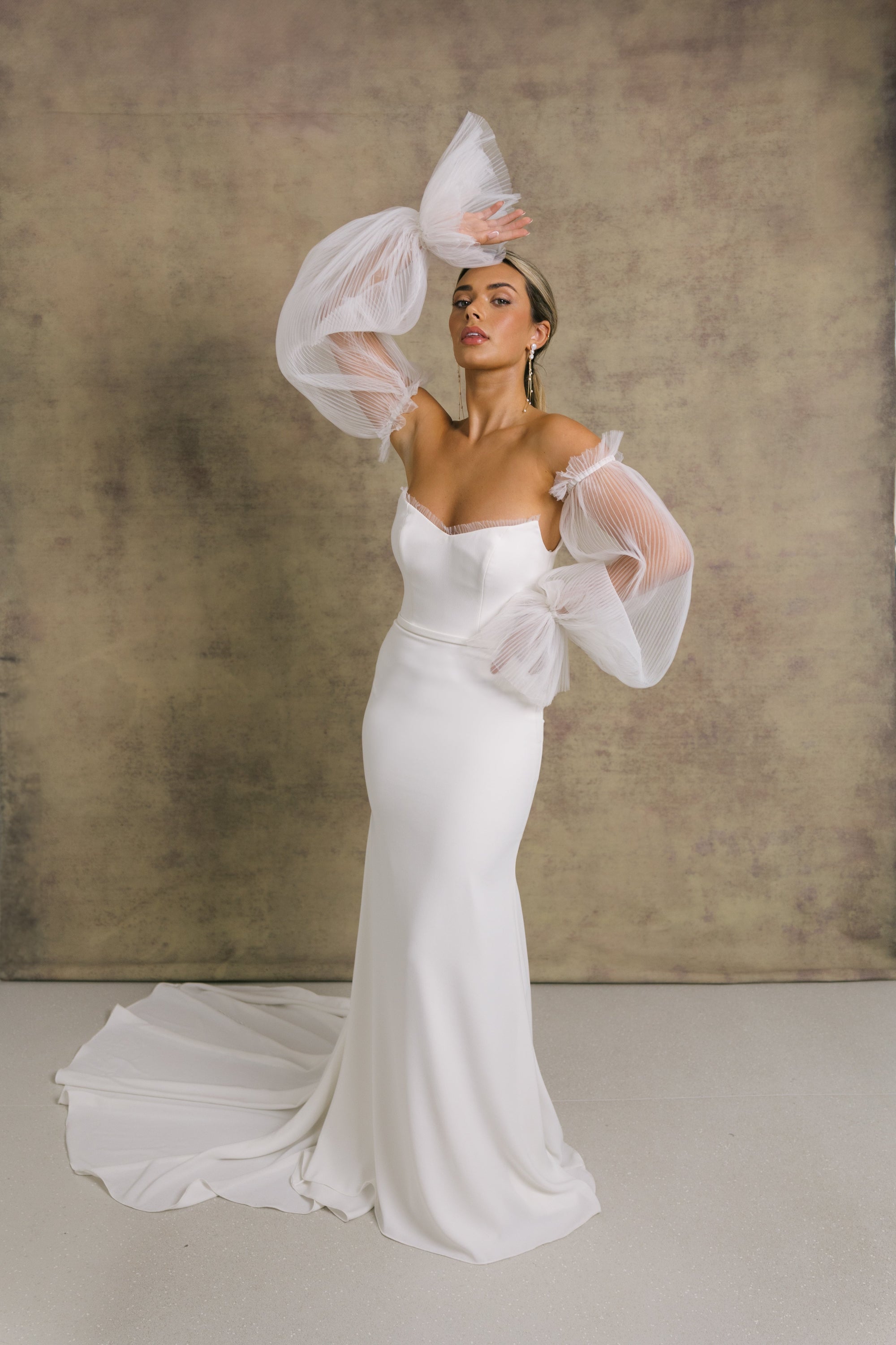 A woman in a strapless ivory wedding gown with a sweetheart neckline and sheer, pleated tulle sleeves showcasing the elegant fit and flare crepe skirt.