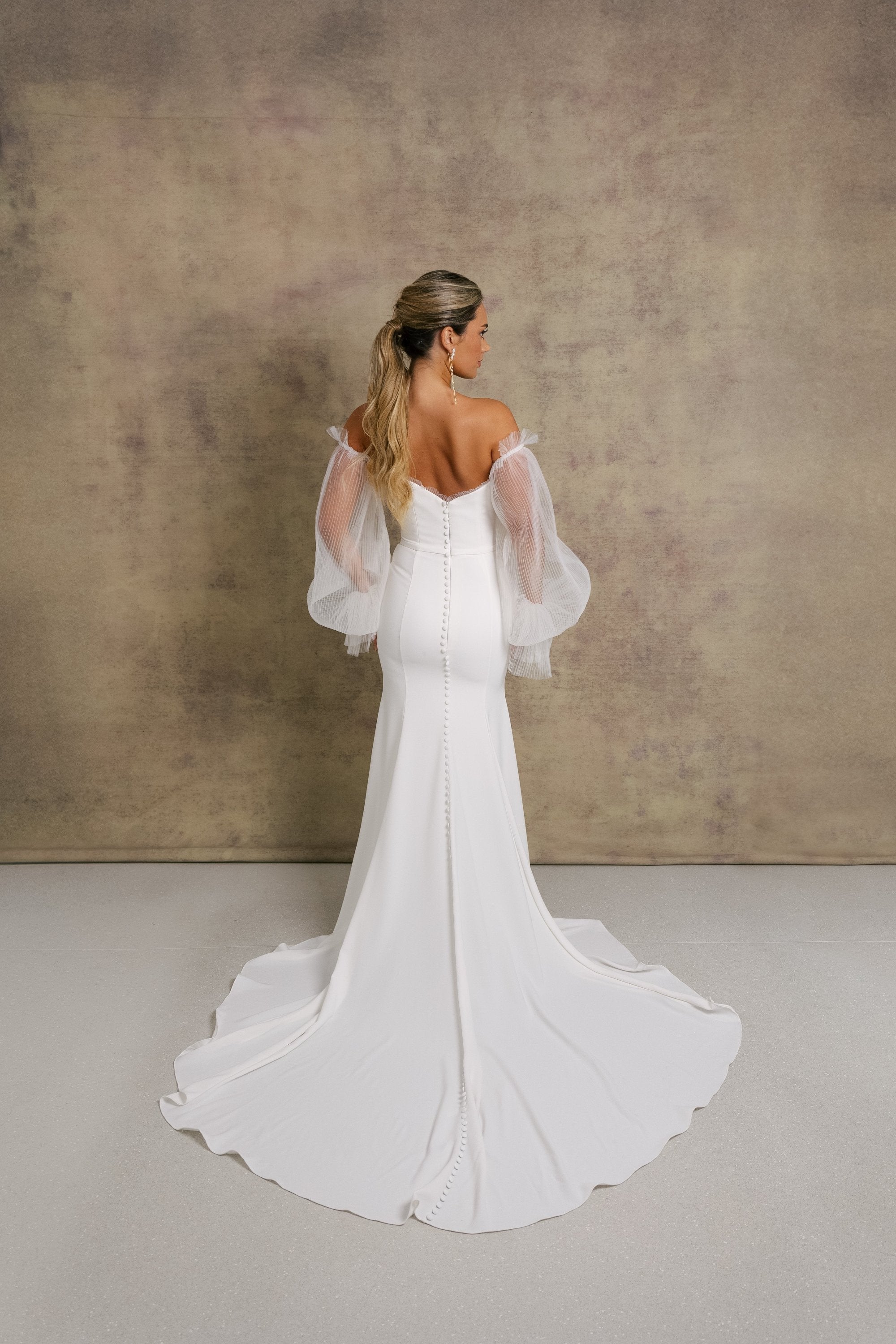 A woman in a strapless ivory wedding gown with a sweetheart neckline and sheer, pleated tulle sleeves showcasing the elegant fit and flare crepe skirt.