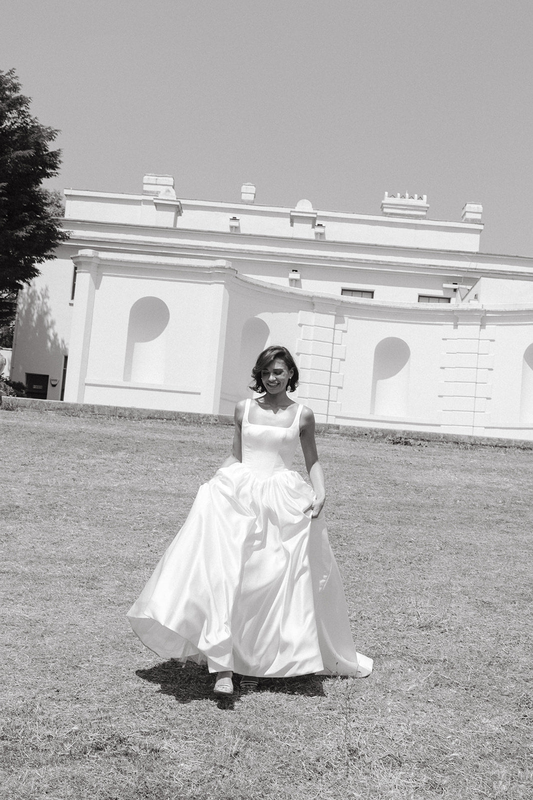 Woman in a ivory satin wedding dress standing on grass with a building in the background