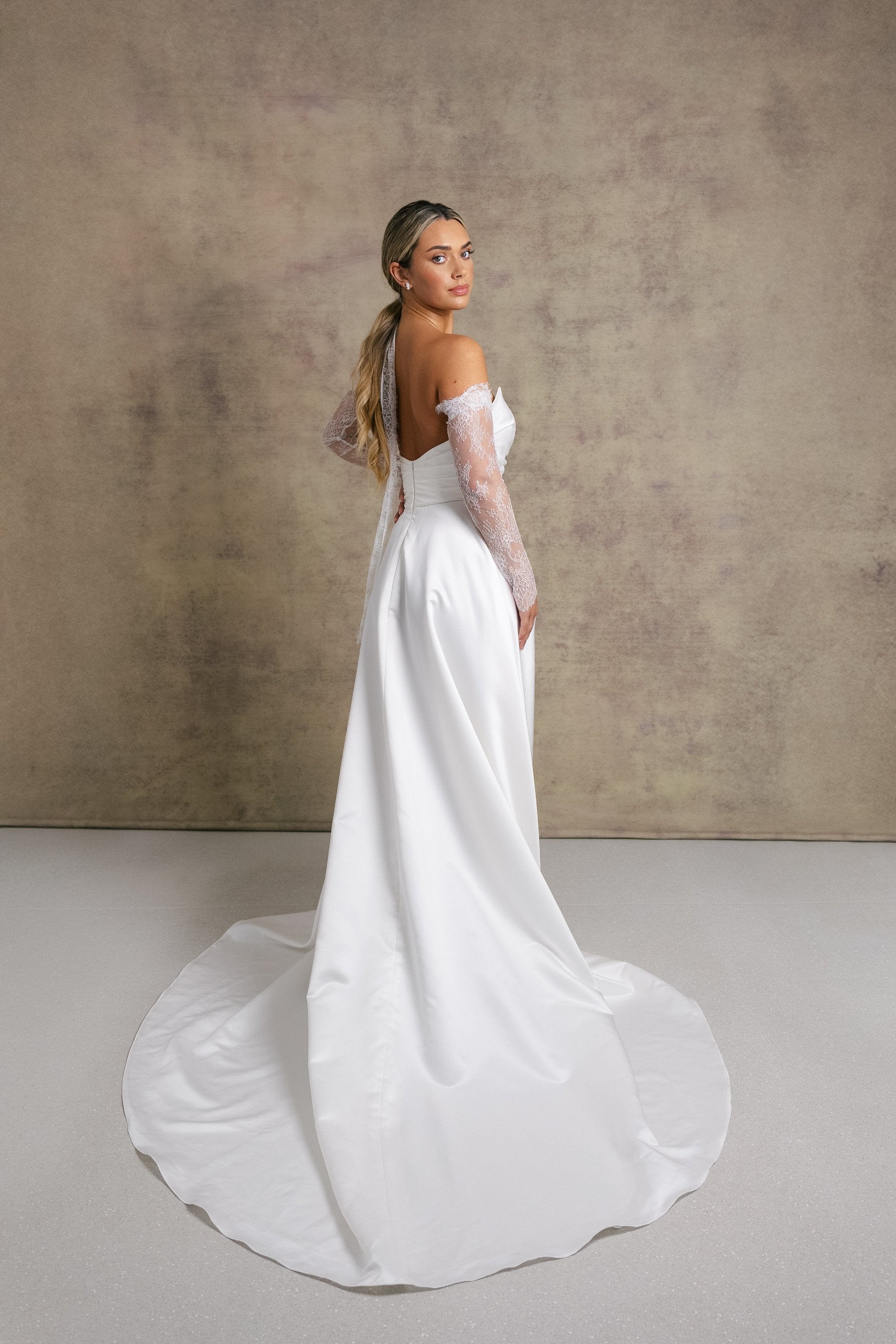 A woman in a satin ivory wedding dress. The strapless dress features a sweetheart neckline and high slit, complemented by chantilly lace sleeves and a matching necktie. 