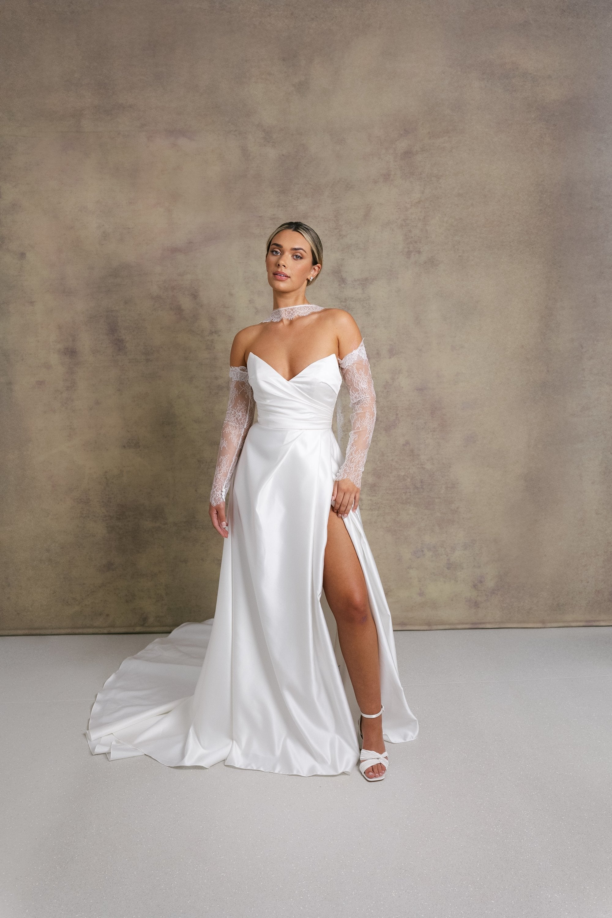 A woman in a satin ivory a line wedding dress. The strapless dress features a sweetheart neckline and high slit, complemented by chantilly lace sleeves and a matching necktie. 