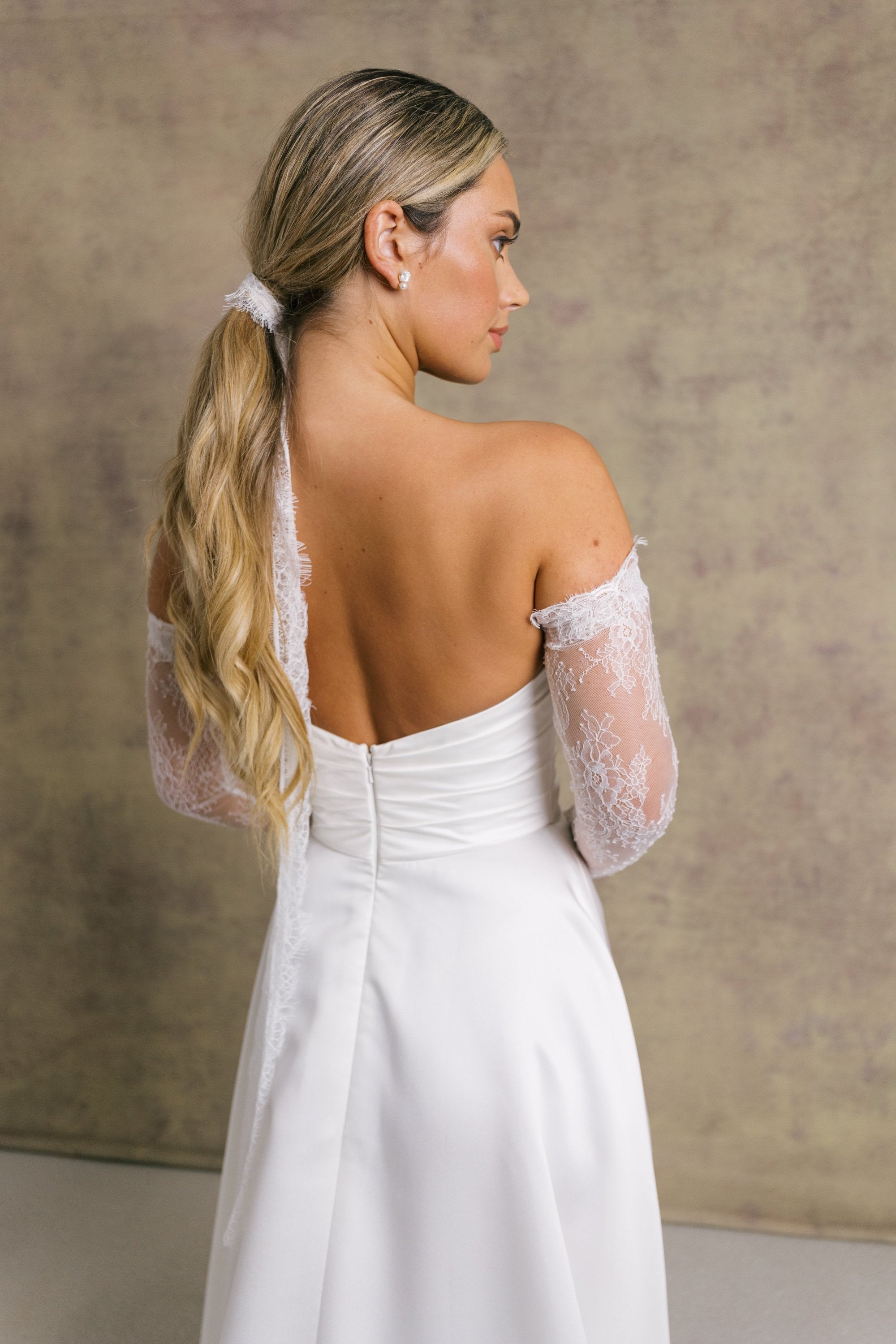 A woman in a satin ivory wedding dress. The strapless dress features a sweetheart neckline and high slit, complemented by chantilly lace sleeves and a matching necktie. 