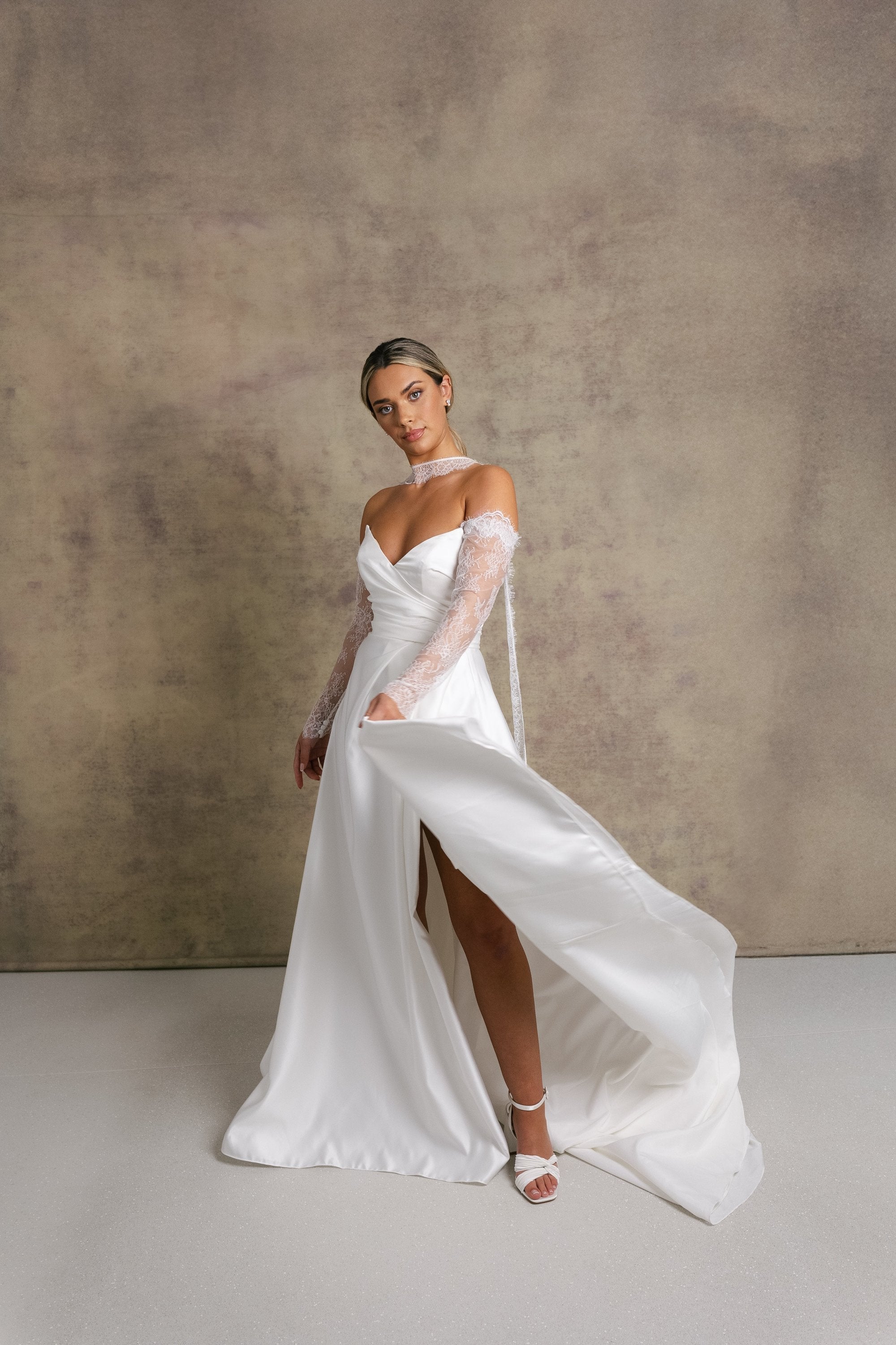 A woman in a satin ivory a line wedding dress. The strapless dress features a sweetheart neckline and high slit, complemented by chantilly lace sleeves and a matching necktie. 