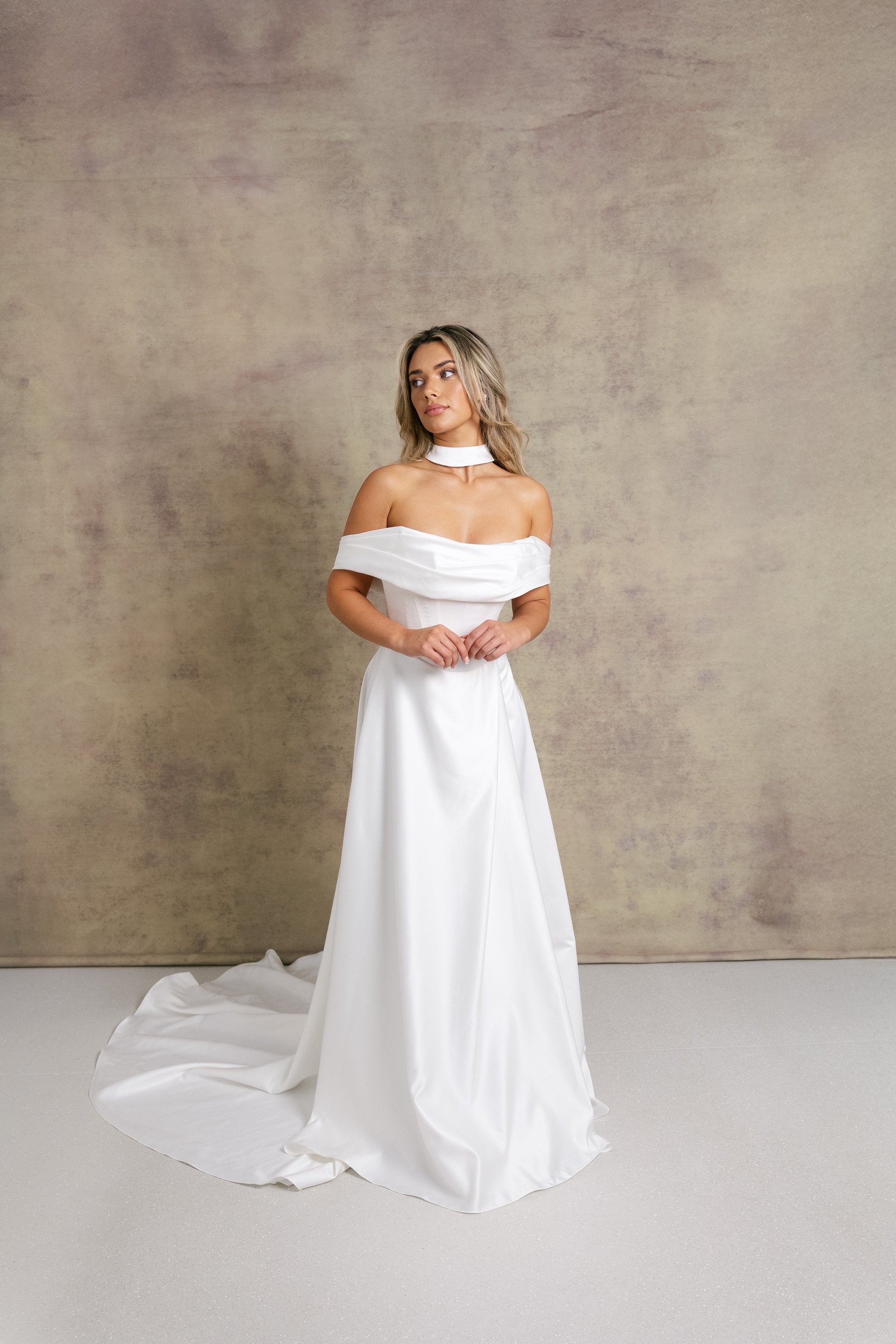 A woman in a subtle dropped waist wedding dress, off the shoulder boned bodice with a soft draped neckline and a wrap over leg split skirt in our gorgeous satin fabric.