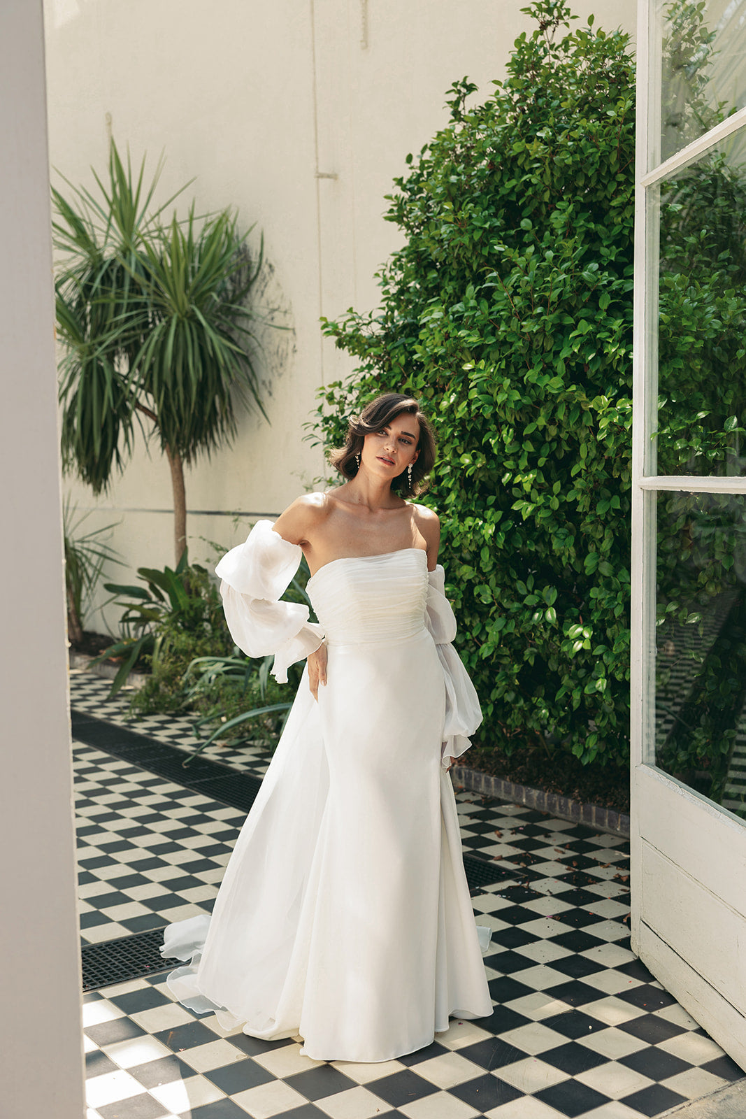 Woman in a ivory wedding dress standing in an outdoor setting with plants and a checkered floor.