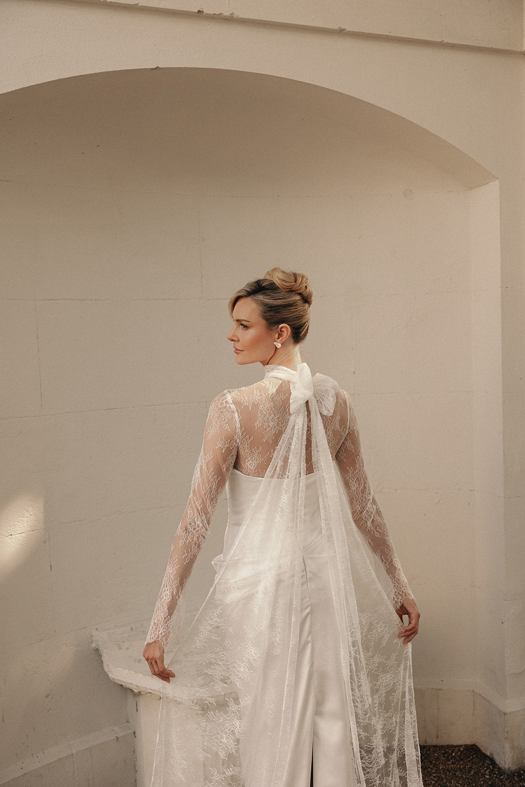 Woman in a ivory wedding dress with lace toppper standing against a plain wall