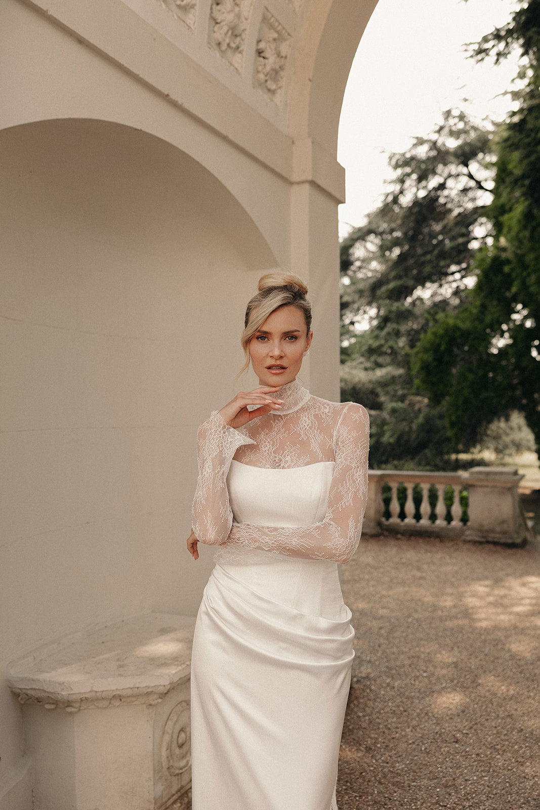 Woman in a ivory wedding lace dress standing in an outdoor setting with classical architecture.