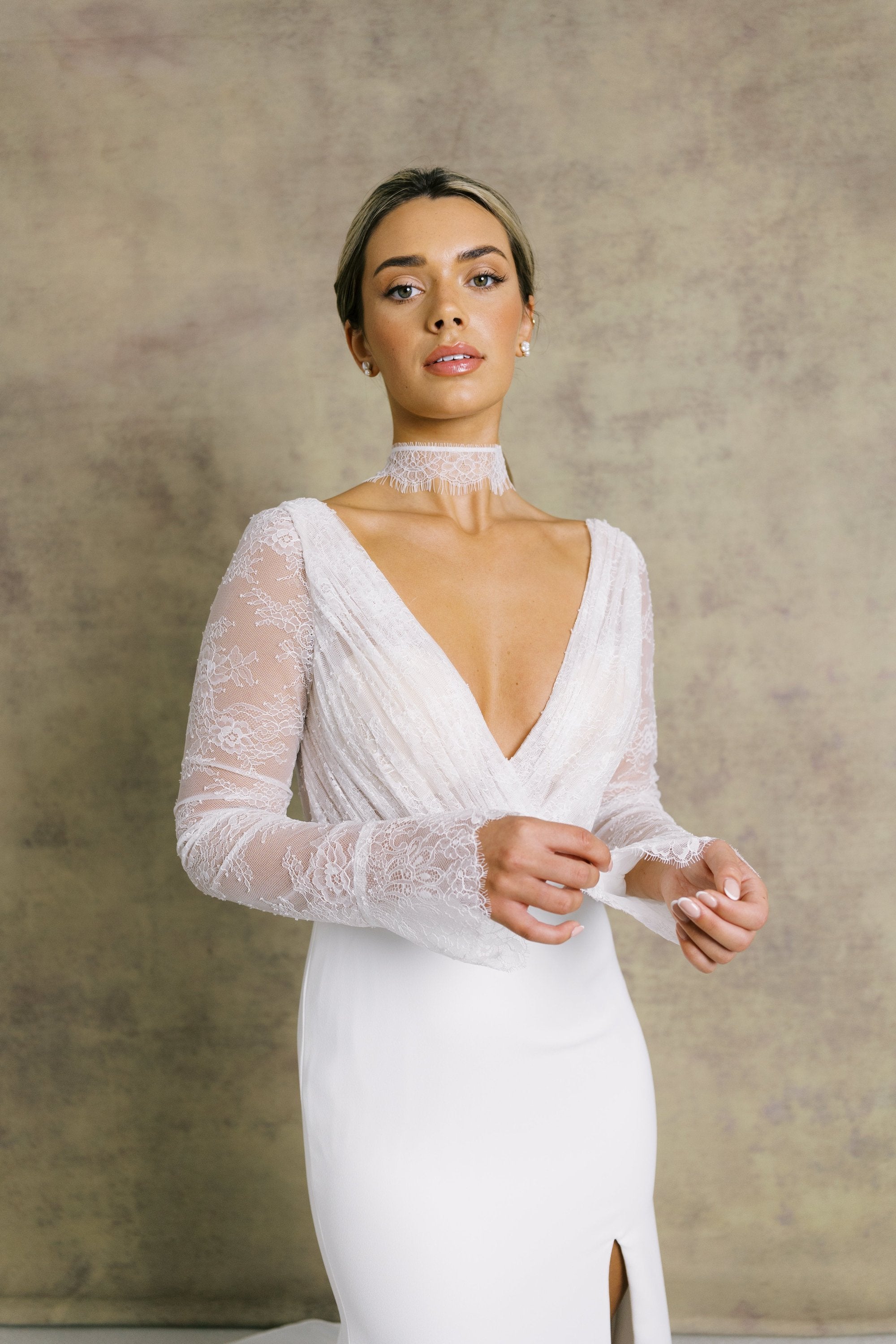 A woman in a pleated chantilly lace v neck bodice wedding dress with long sleeves, finished with dreamy, flared cuffs, complemented on fitted crepe skirt.