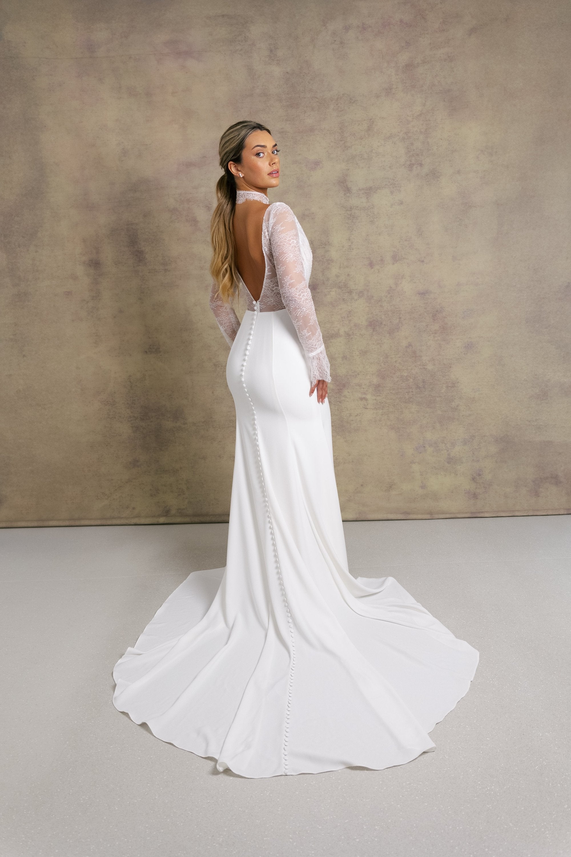 A woman in a pleated chantilly lace v neck bodice wedding dress with long sleeves, finished with dreamy, flared cuffs, complemented on fitted crepe skirt.