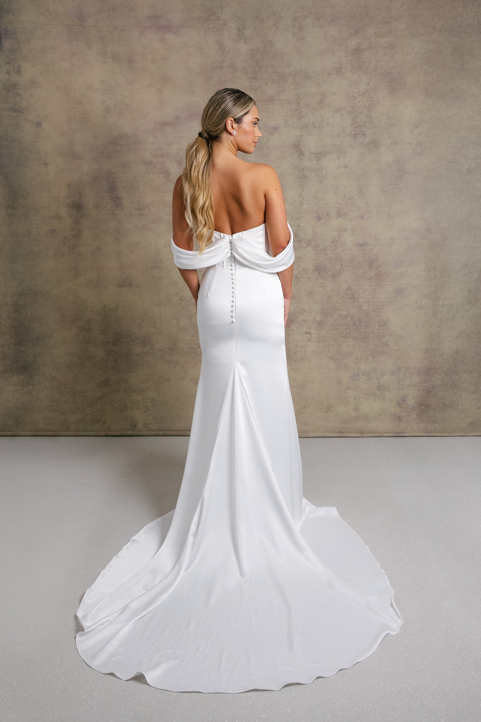 A woman in luxurious slipper satin wedding dress featuring a draped neckline on an exposed boned corset style drop waist bodice with draped bardot’s on our new ruched skirt.