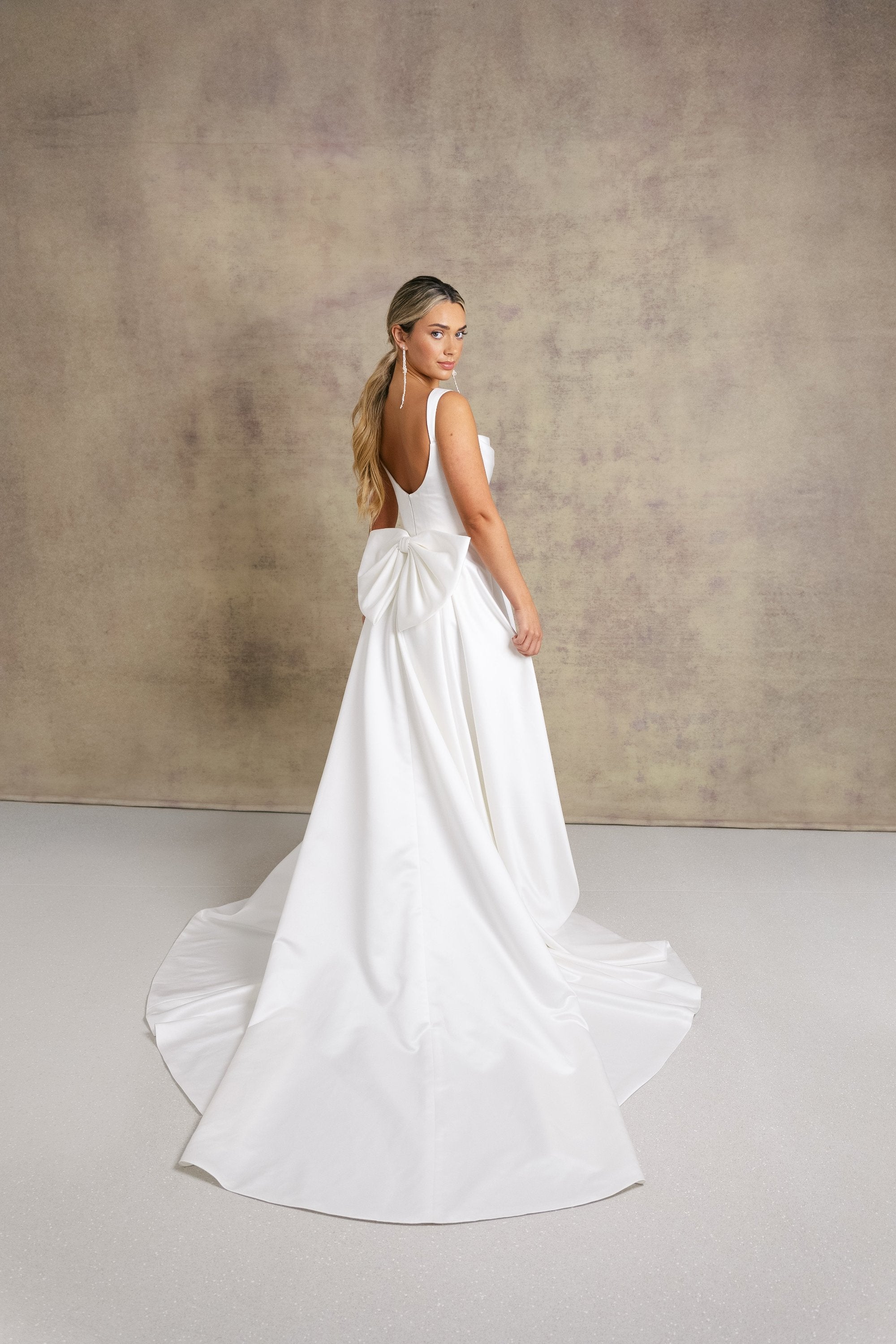 A woman in a satin striking cowl neckline wedding dress with a box pleated soft Aline skirt and bow.