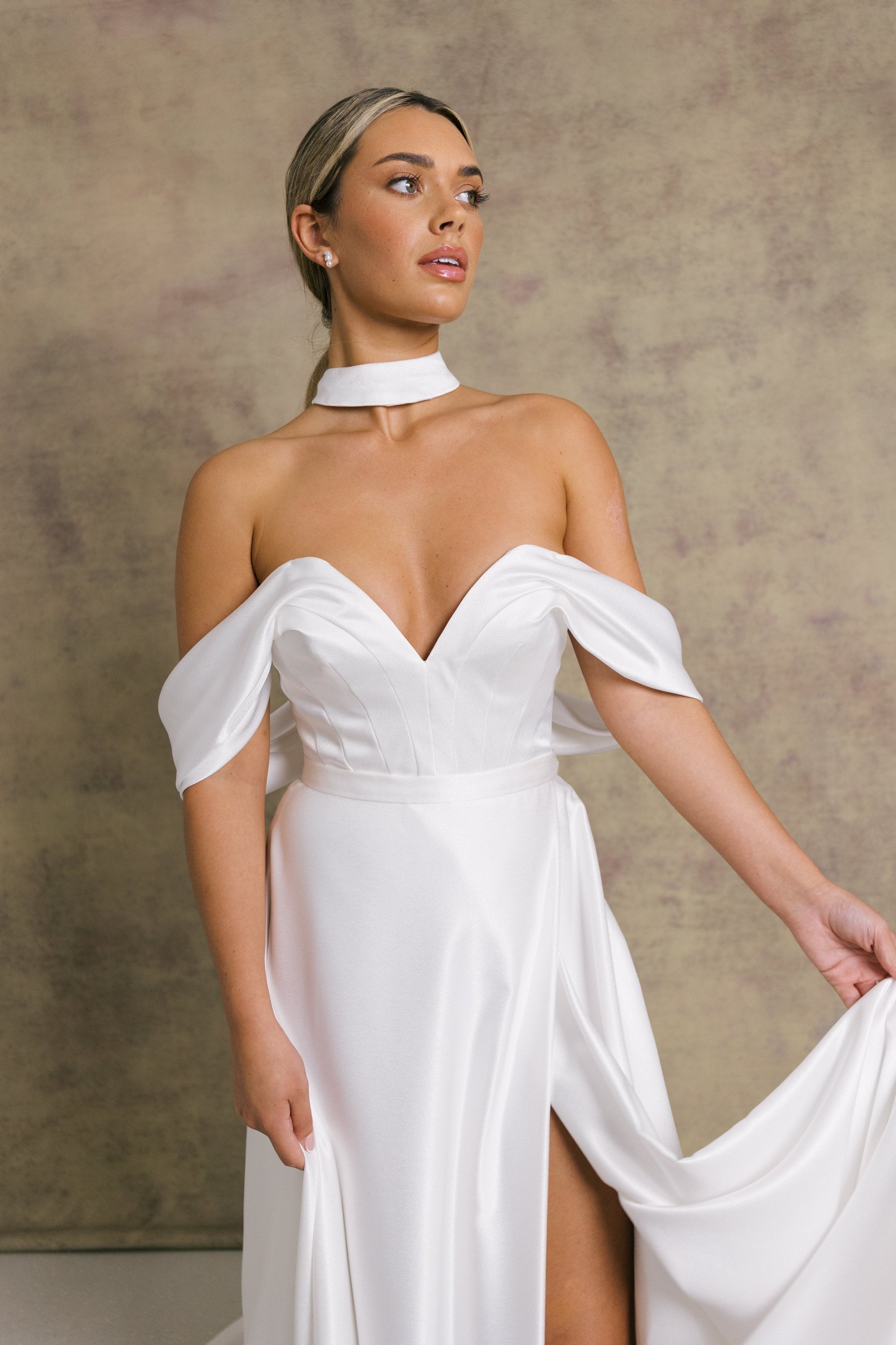 A woman in a luxurious slipper satin two piece wedding dress, featuring a sweetheart corset top with off the shoulder bardot sleeves and slipper satin wrap over leg split skirt.