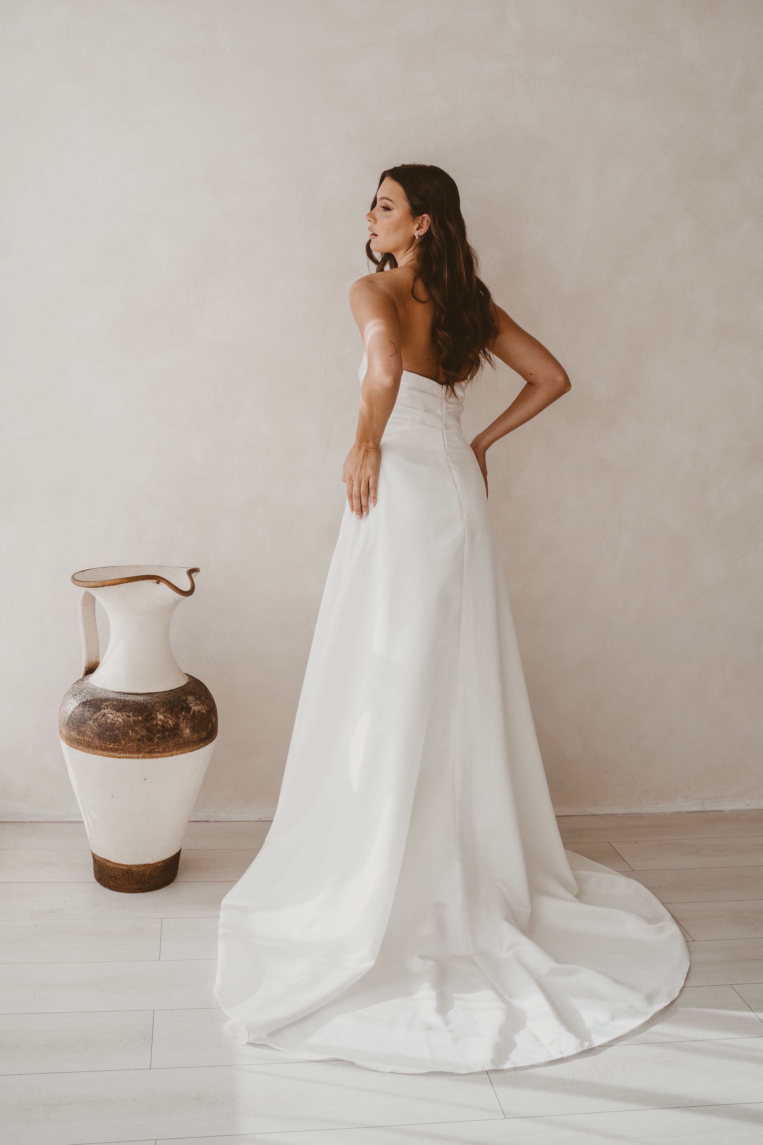 Woman in a white strapless gown standing in a minimalistic room with a vase.