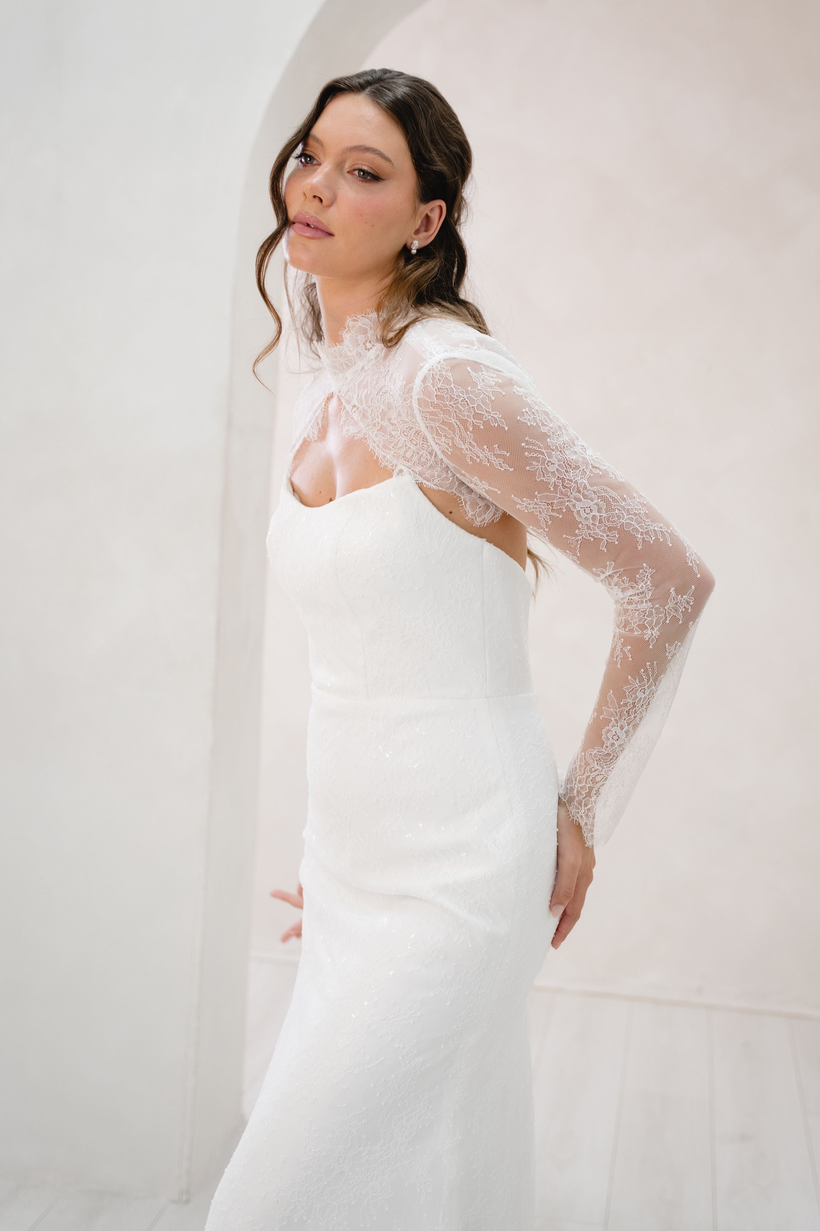 Woman wearing a white wedding dress with sheer sleeves against a plain background