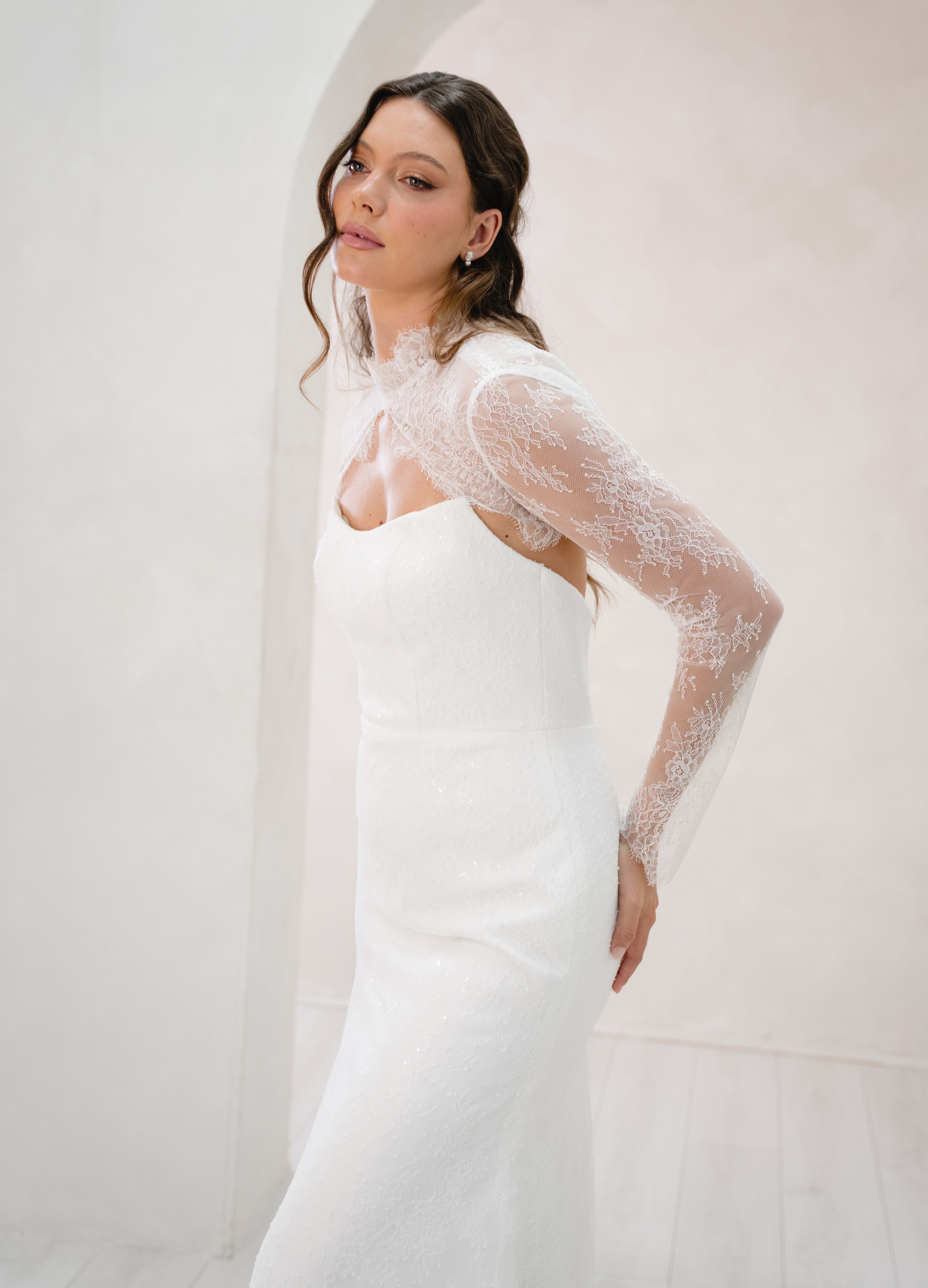 Woman wearing a white wedding dress with sheer sleeves against a plain background