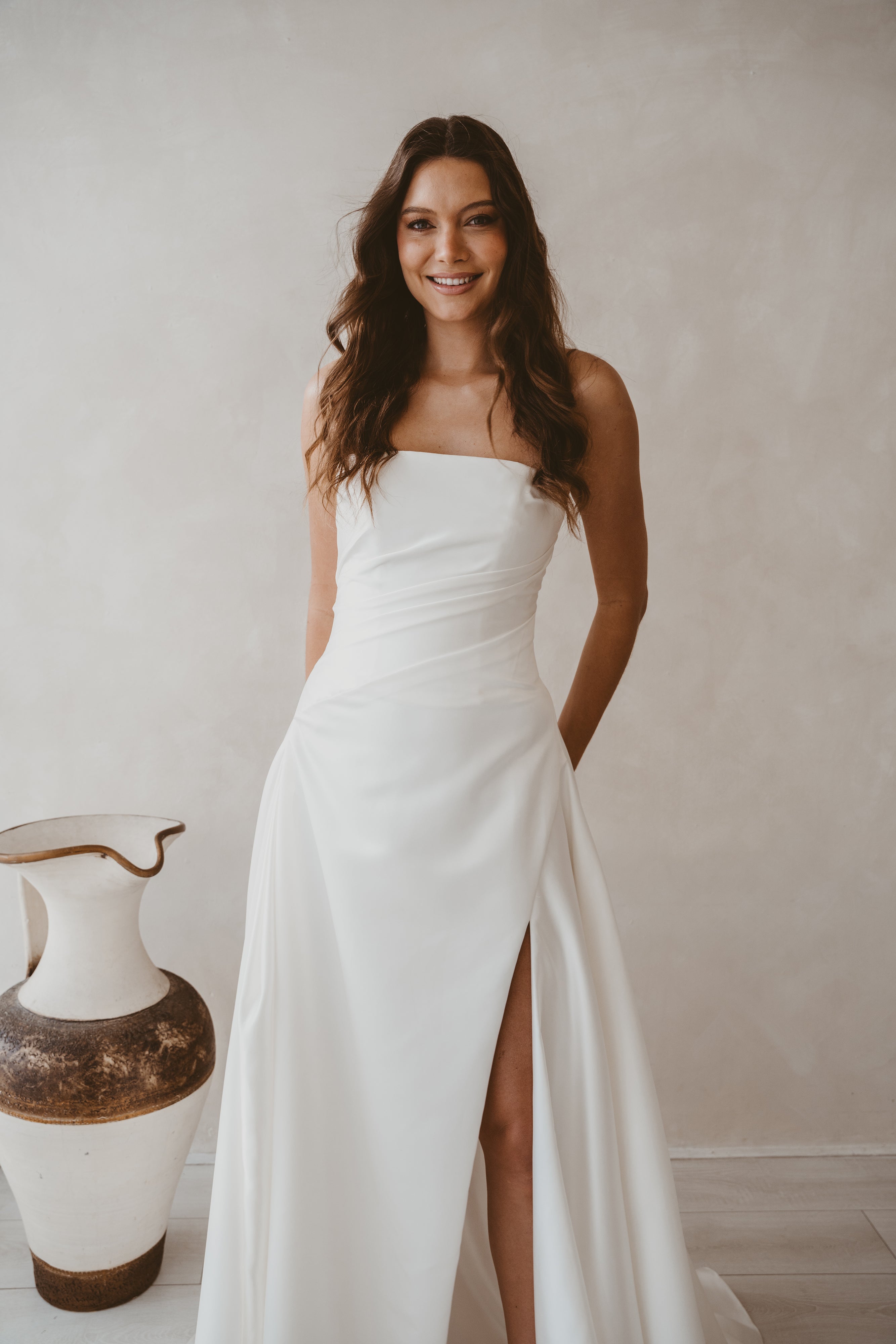 Woman in a white strapless gown with a slit standing against a plain wall.