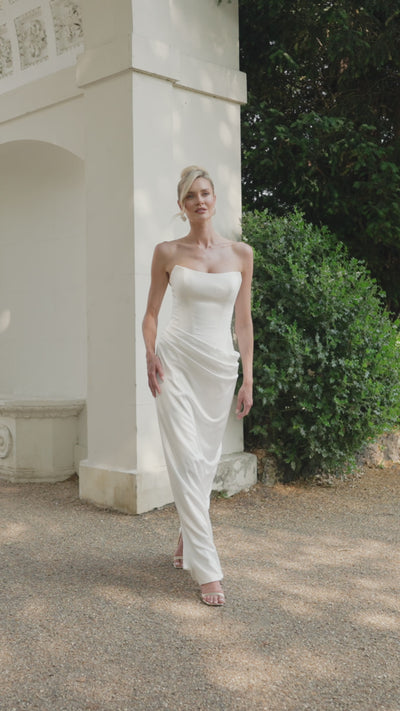 Woman in a strapless dropped waist wedding dress moving next to a classical architectural structure in a garden.