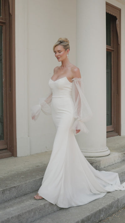 A woman in a strapless ivory wedding gown with a sweetheart neckline and sheer, pleated tulle sleeves showcasing the elegant fit and flare crepe skirt.