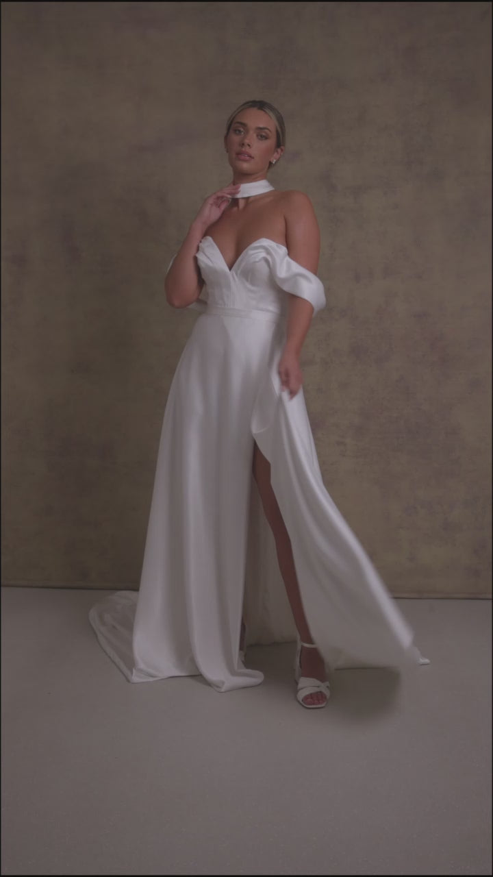 A woman in a luxurious slipper satin two piece wedding dress, featuring a sweetheart corset top with off the shoulder bardot sleeves and slipper satin wrap over leg split skirt.