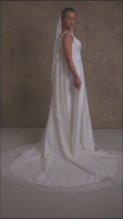 A woman in a striking v neckline wedding dress with waistband detail on a box pleated satin skirt, complemented with a Chantilly lace border veil.