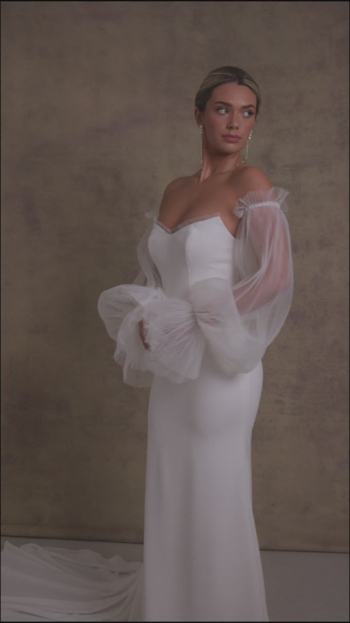 A woman in a strapless ivory wedding gown with a sweetheart neckline and sheer, pleated tulle sleeves showcasing the elegant fit and flare crepe skirt.