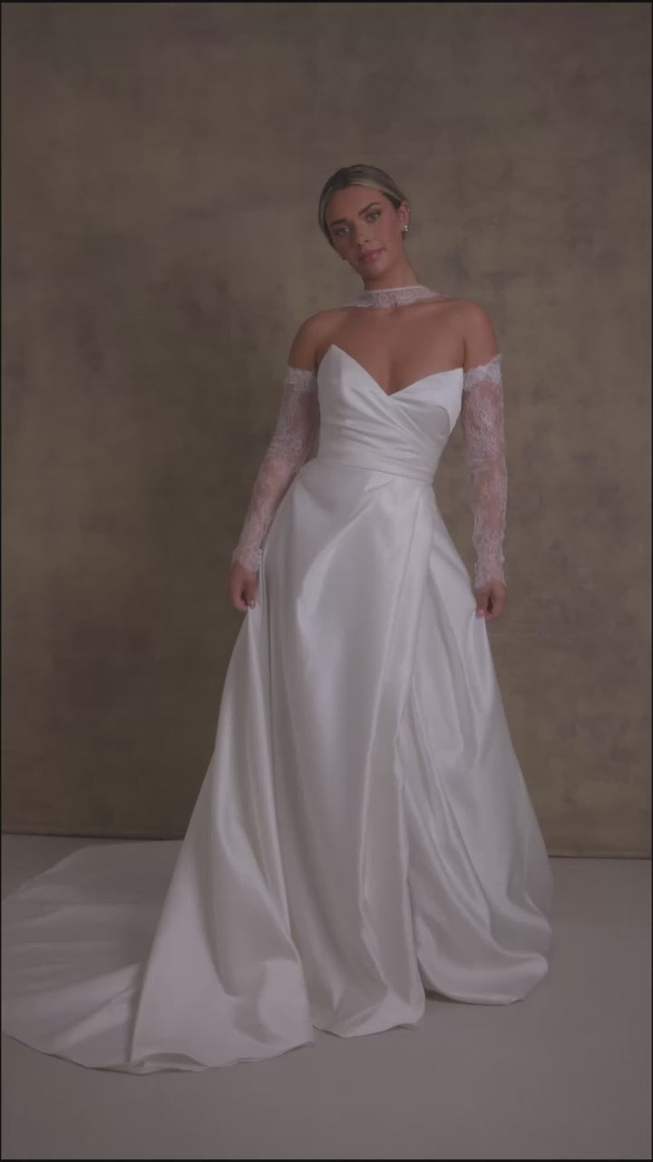 A woman in a satin ivory a line wedding dress. The strapless dress features a sweetheart neckline and high slit, complemented by chantilly lace sleeves and a matching necktie. 