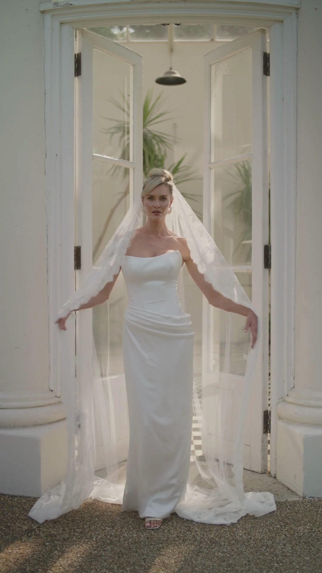 Woman in a ivory wedding dress and long lace wedding veil moving in front of a large glass door with columns.