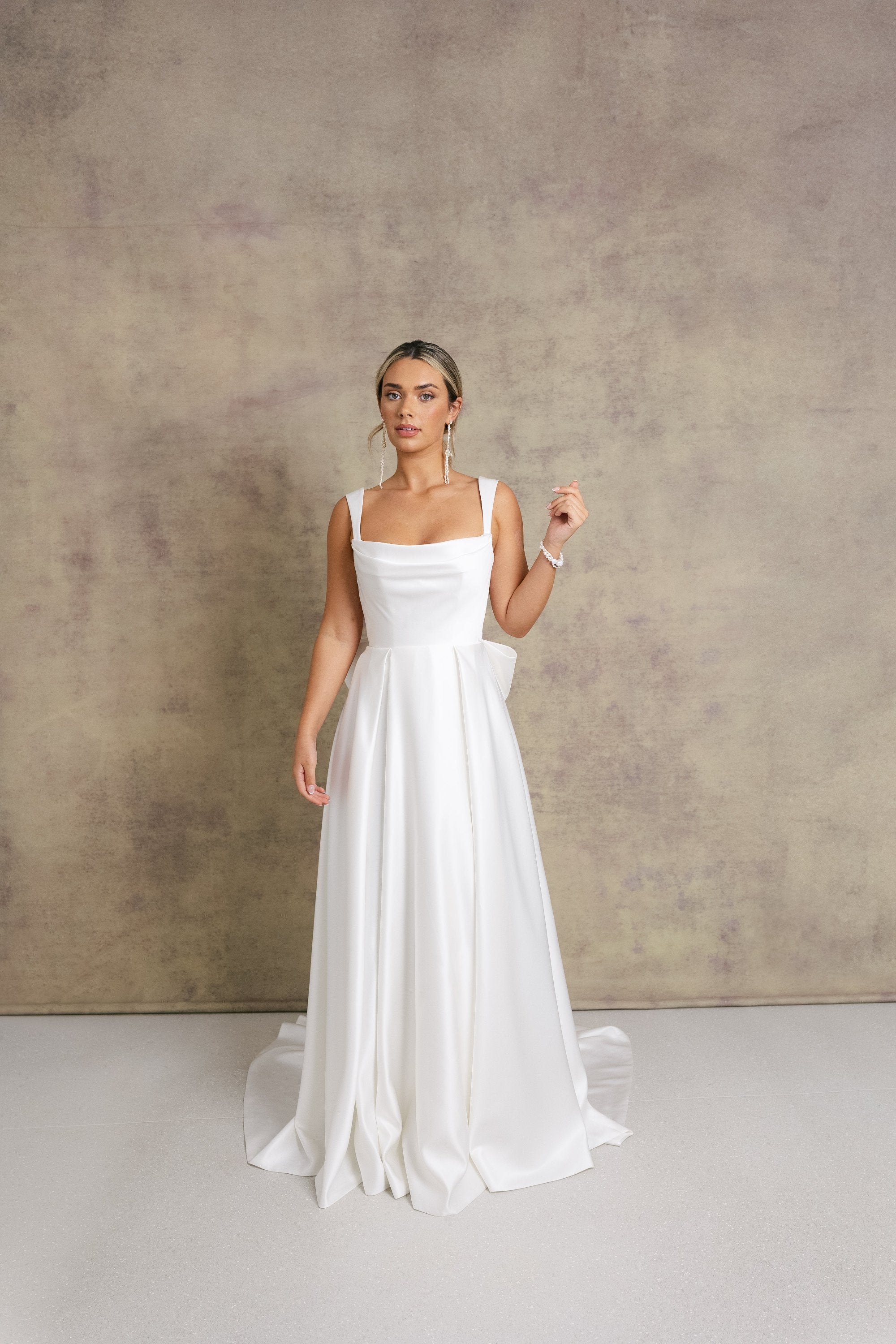 A woman in a satin striking cowl neckline wedding dress with a box pleated soft Aline skirt and bow.