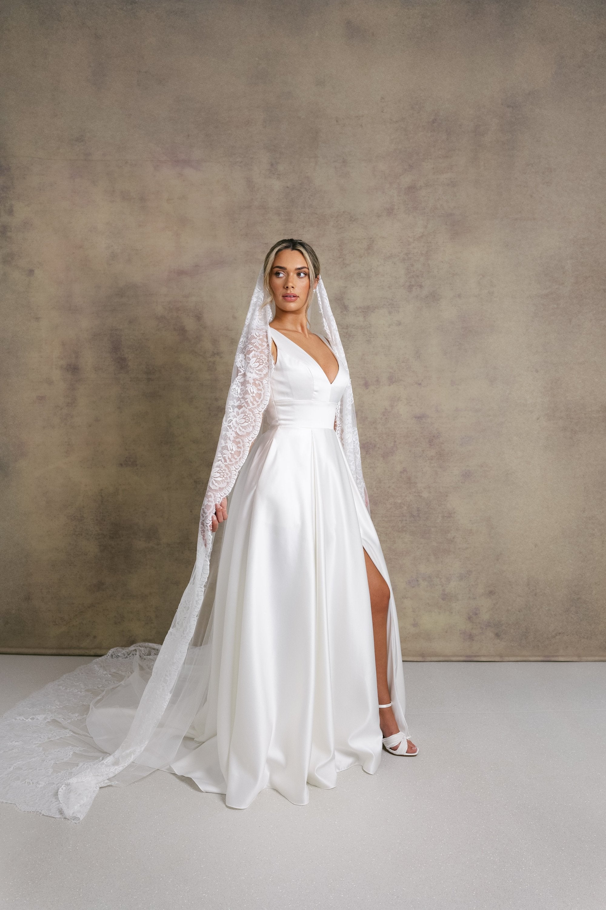 A woman in a striking v neckline wedding dress with waistband detail on a box pleated satin skirt, complemented with a Chantilly lace border veil.