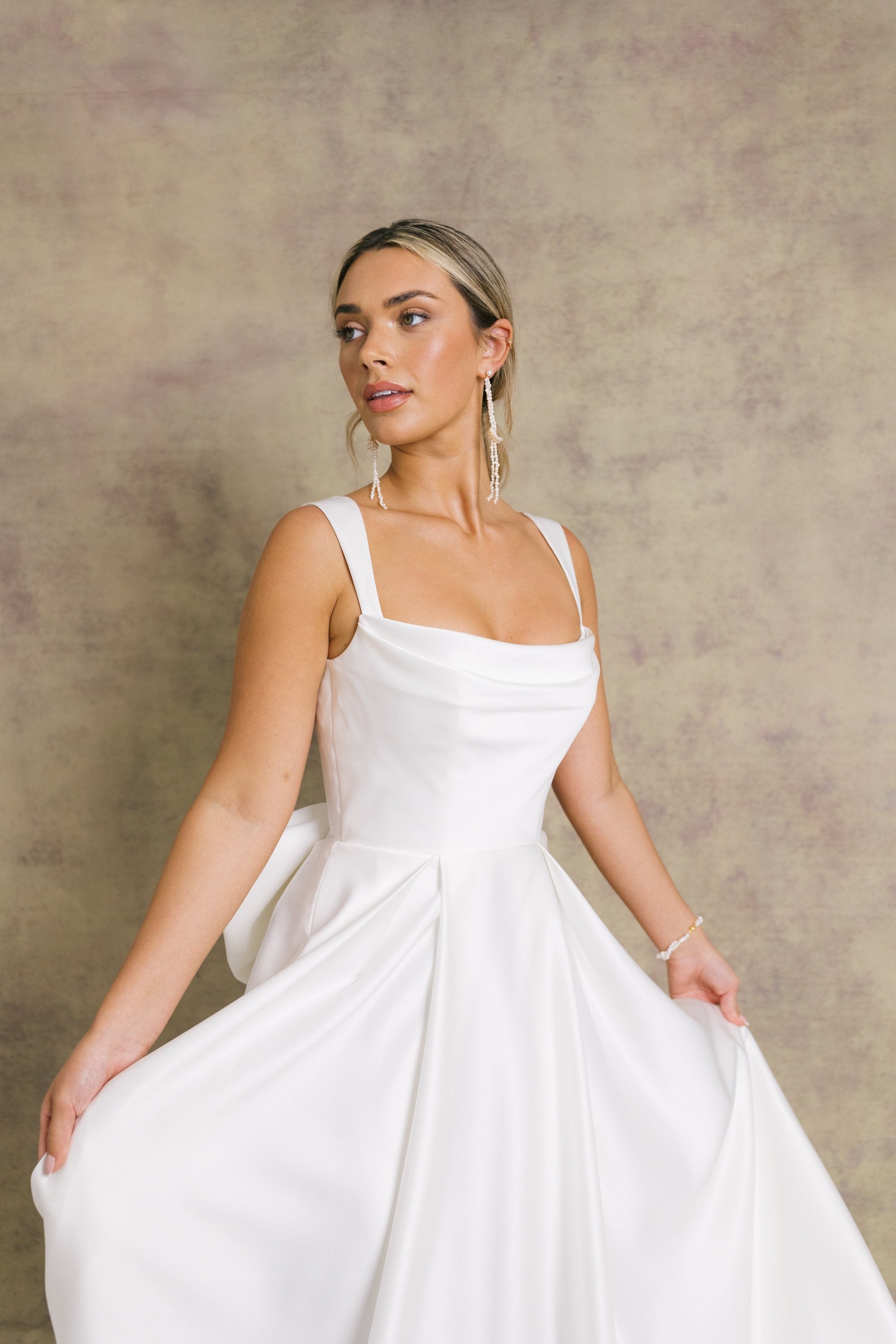 A woman in a satin striking cowl neckline wedding dress with a box pleated soft Aline skirt and bow.