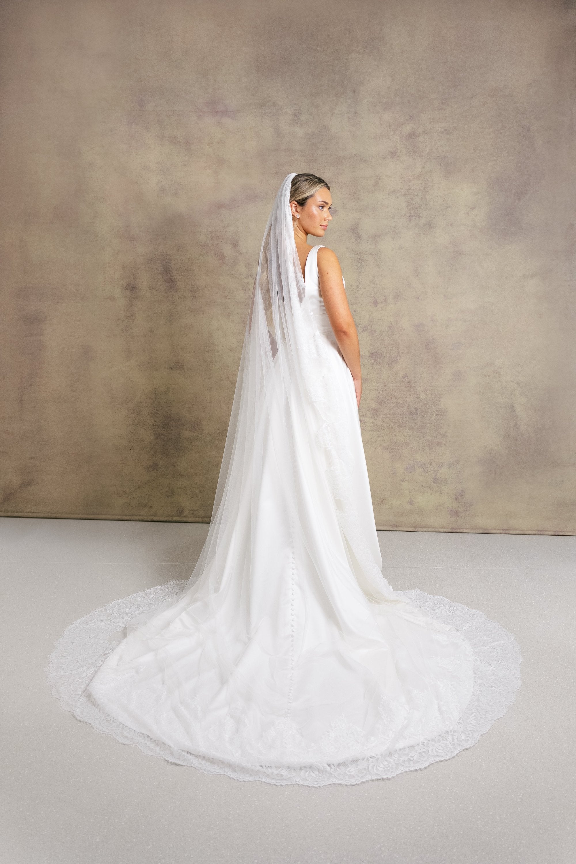 A woman in a striking v neckline wedding dress with waistband detail on a box pleated satin skirt, complemented with a Chantilly lace border veil.