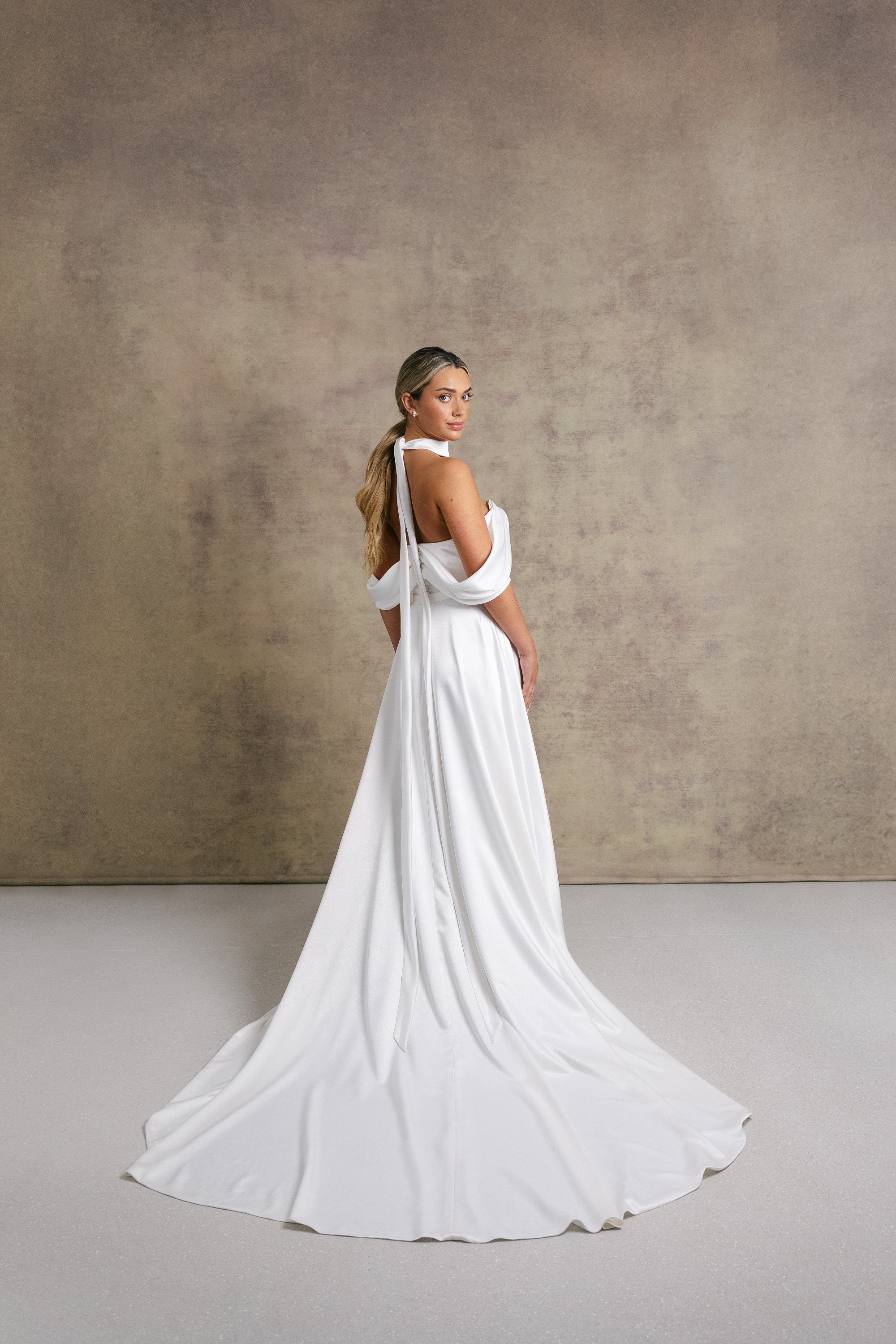 A woman in a luxurious slipper satin two piece wedding dress, featuring a sweetheart corset top with off the shoulder bardot sleeves and slipper satin wrap over leg split skirt.